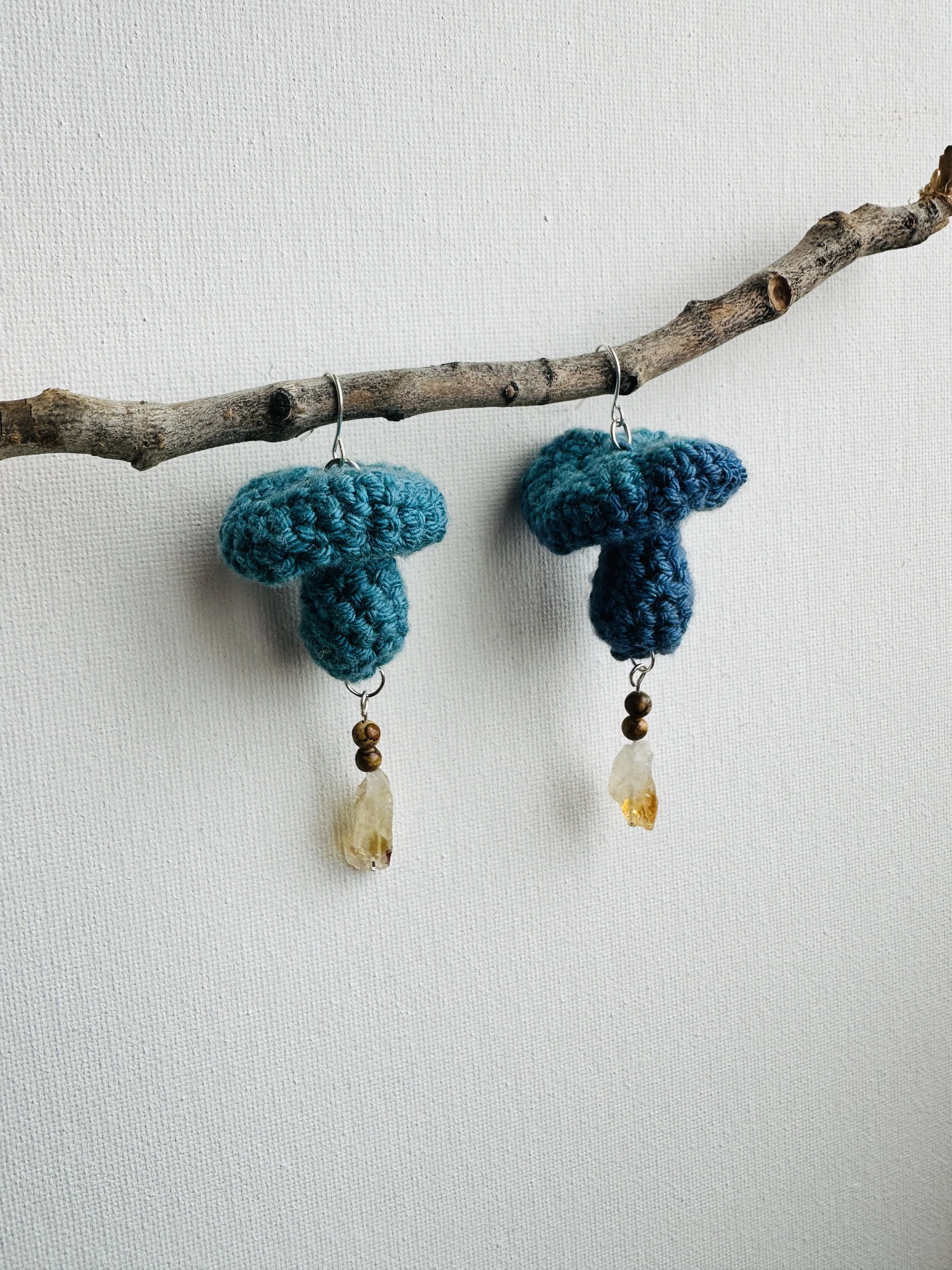 Handmade earrings with blue crocheted mushroom designs, mounted on a thin wire, hanging from a branch, with decorative stone and beads.