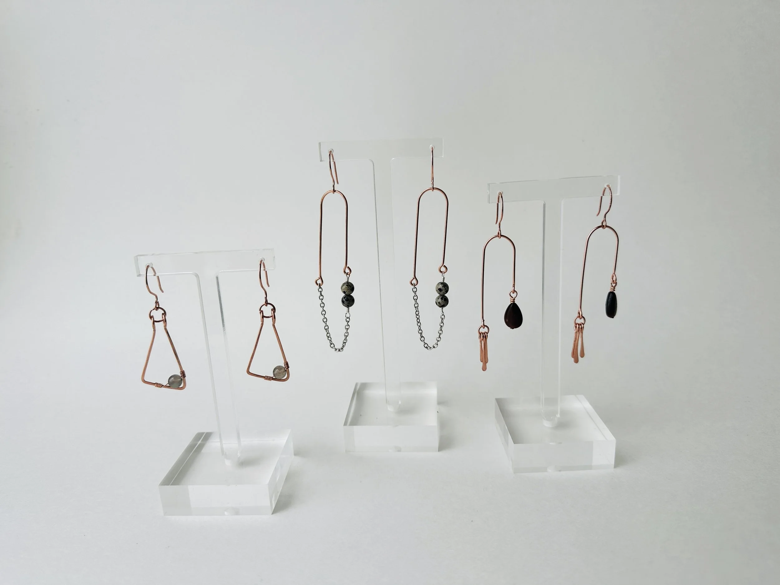 Display of multiple earrings on clear acrylic stands, showcasing various copper, black, and beaded designs against a plain white background.
