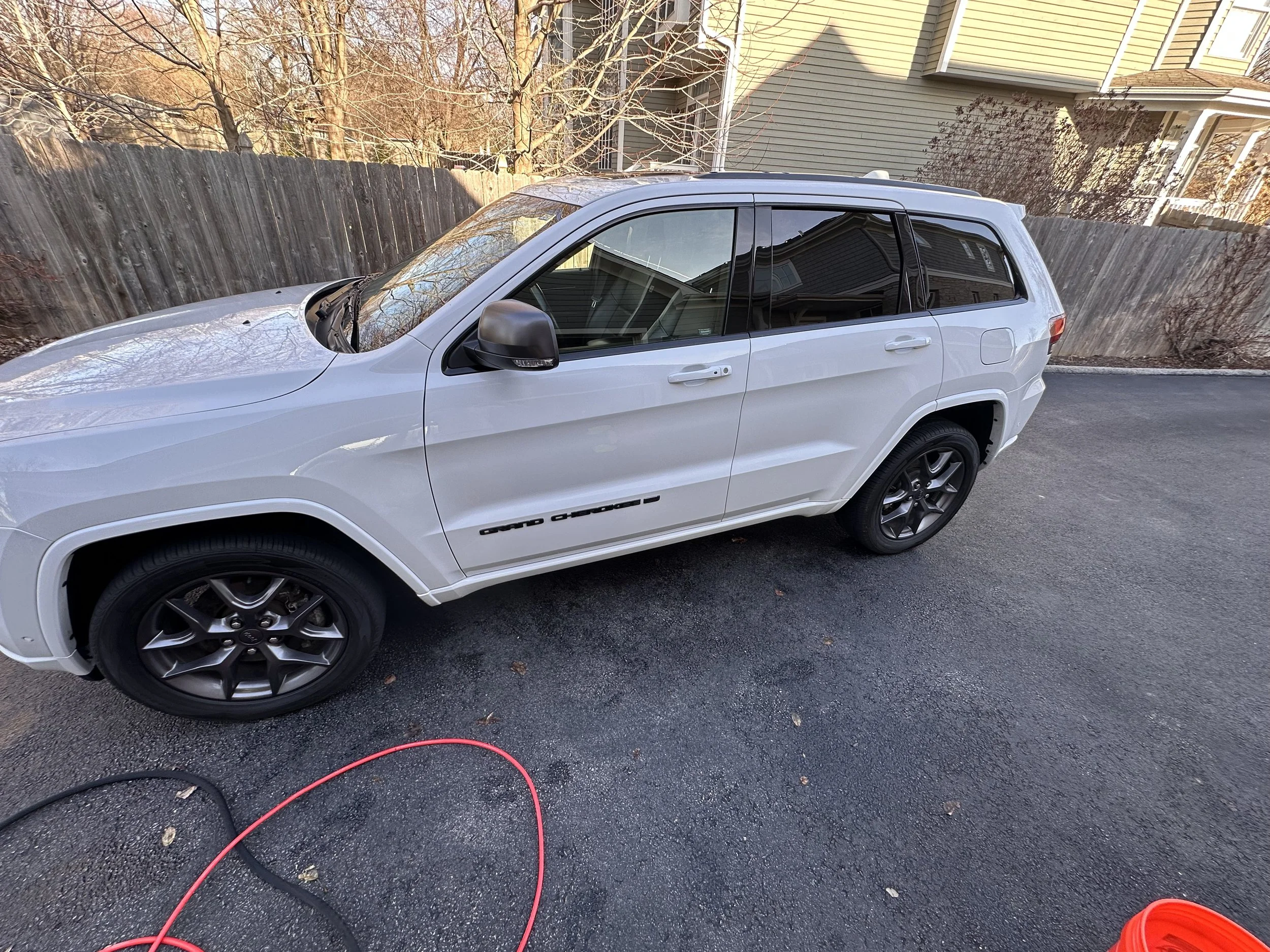 White SUV parked on a driveway with a wooden fence and house in the background, leafless trees, and garden hose on the ground.