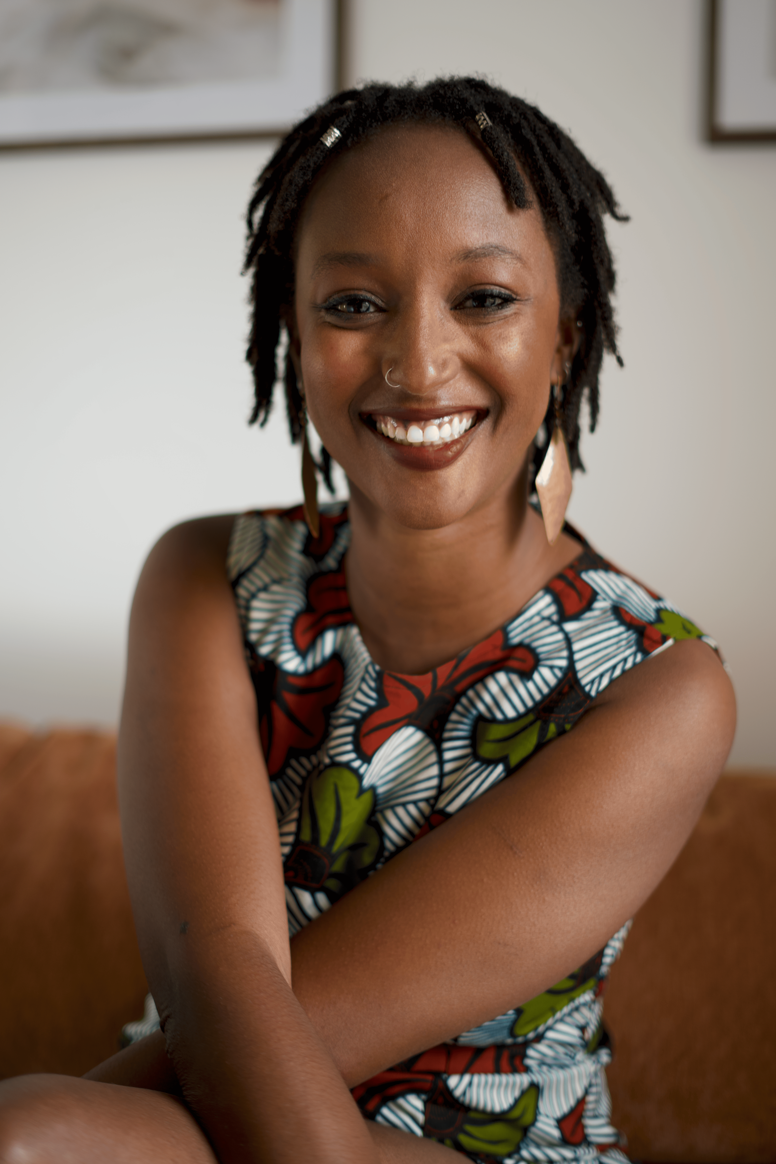 Une femme afro-américaine souriante avec une coiffure en dreadlocks, portant une robe colorée à motifs, assise sur un canapé en cuir marron, dans un intérieur lumineux.