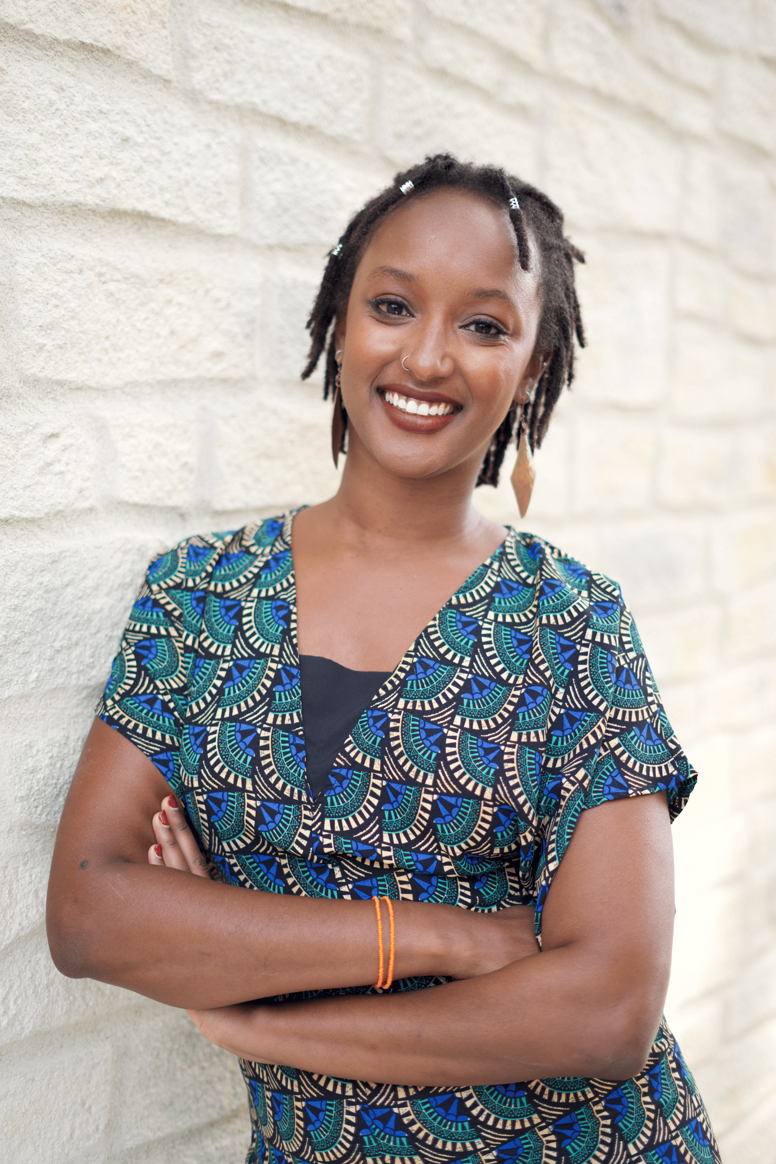 Une femme souriante avec des cheveux en dreadlocks, portant un vêtement à motifs colorés bleus, verts et noirs, avec des boucles d'oreilles et une bague orange, posant contre un mur en pierre beige.