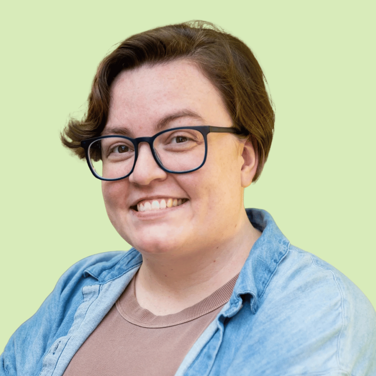 Lillian cotter, Headshot, a woman with short brown hair, glasses, and a big smile wearing a light blue denim shirt over a tan t-shirt, against a light green background.