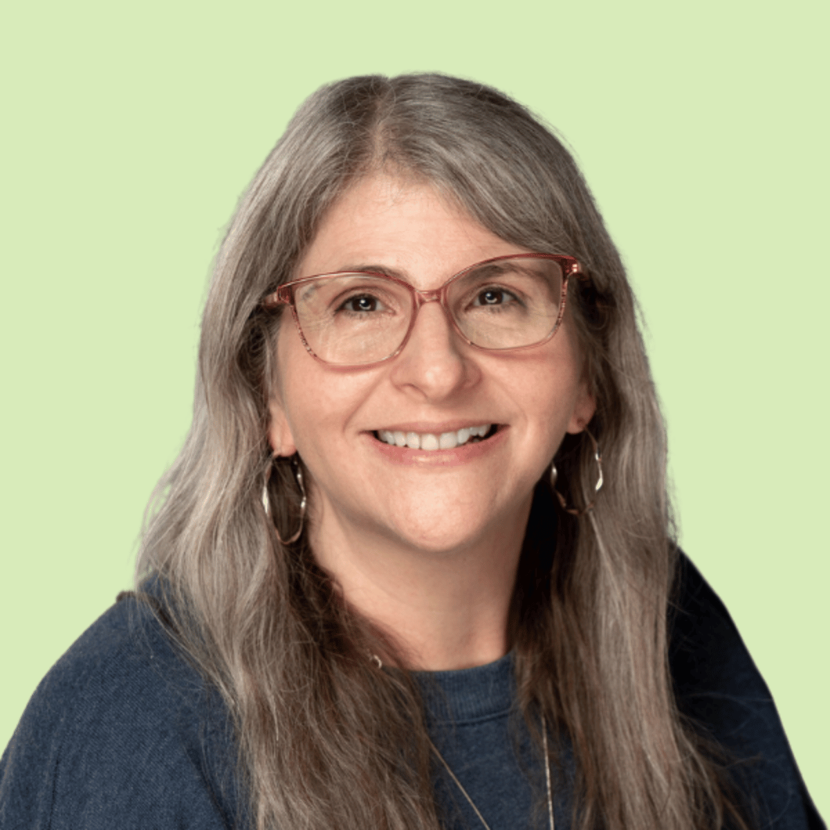 Julie Golden Headshot, A woman with long gray hair, wearing glasses, earrings, and a dark top, smiling against a light green background.
