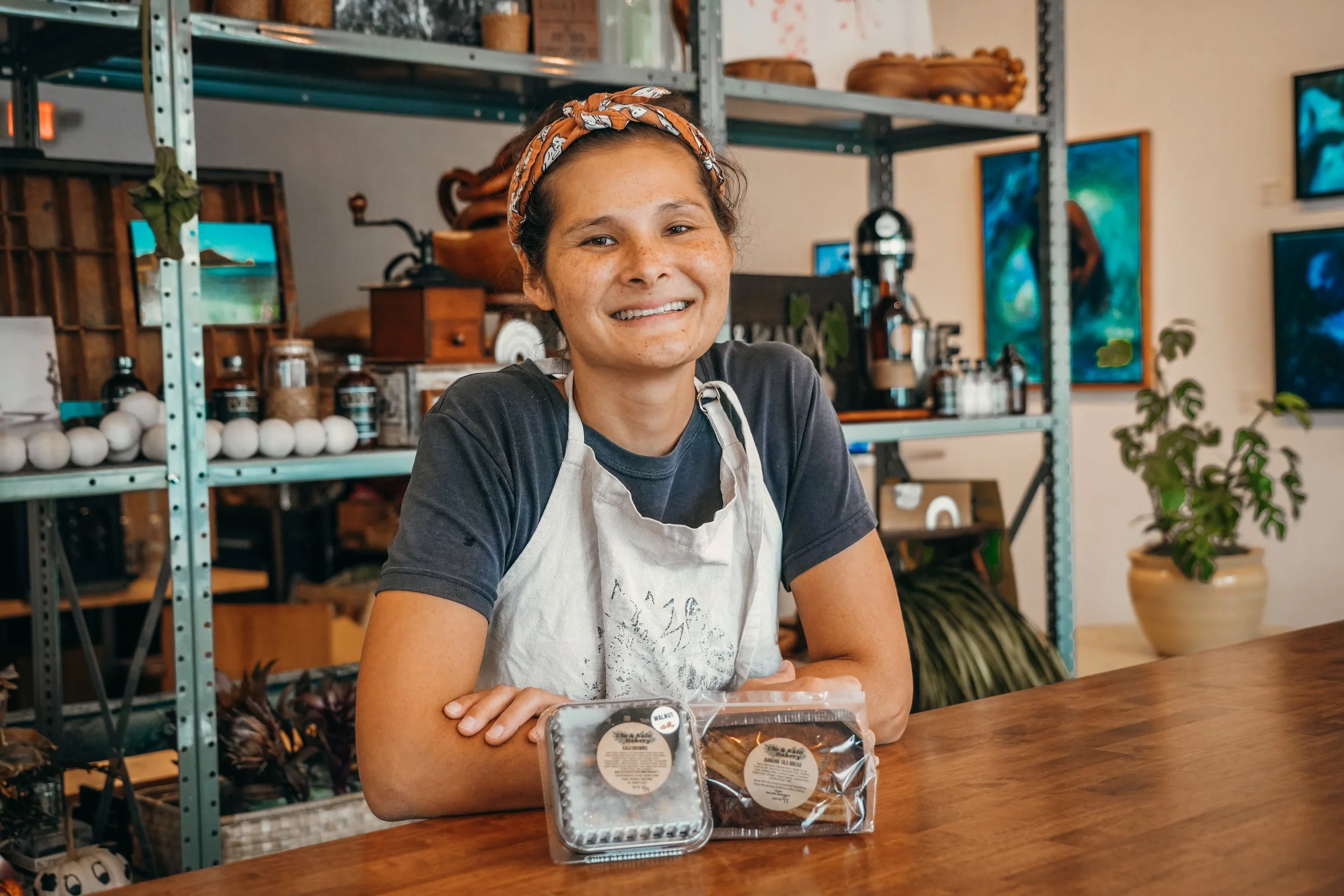 A woman with a headscarf and an apron seated at a wooden table, smiling at the camera. Shelves in the background display various wooden objects, bottles, and decorations, with a potted plant nearby.
