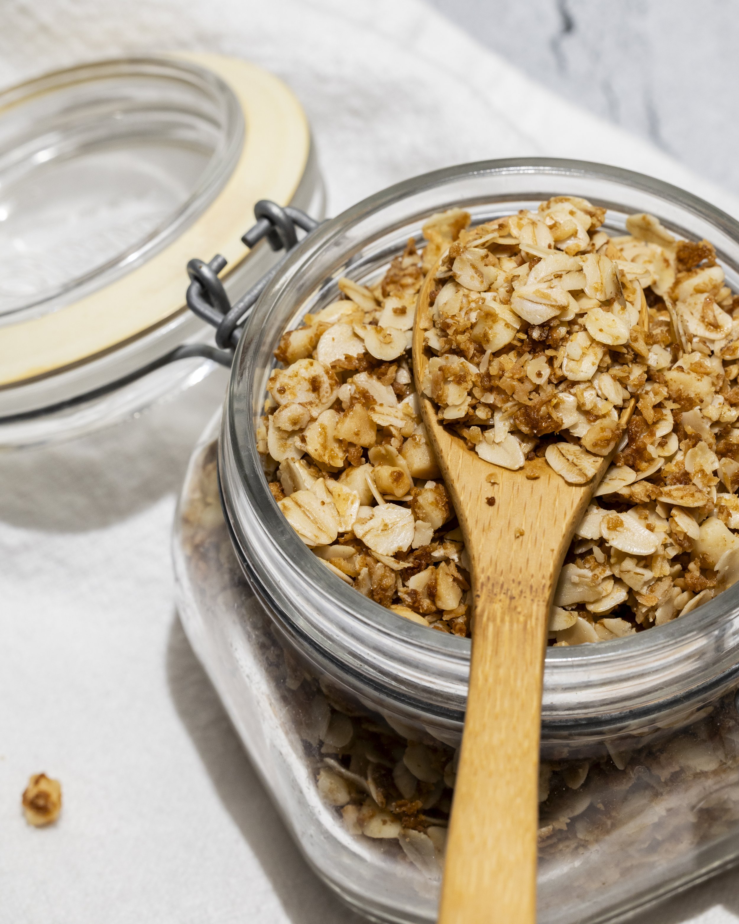 Close-up of a glass jar filled with chopped nuts with a wooden spoon inside, placed on a white surface with some scattered nuts.
