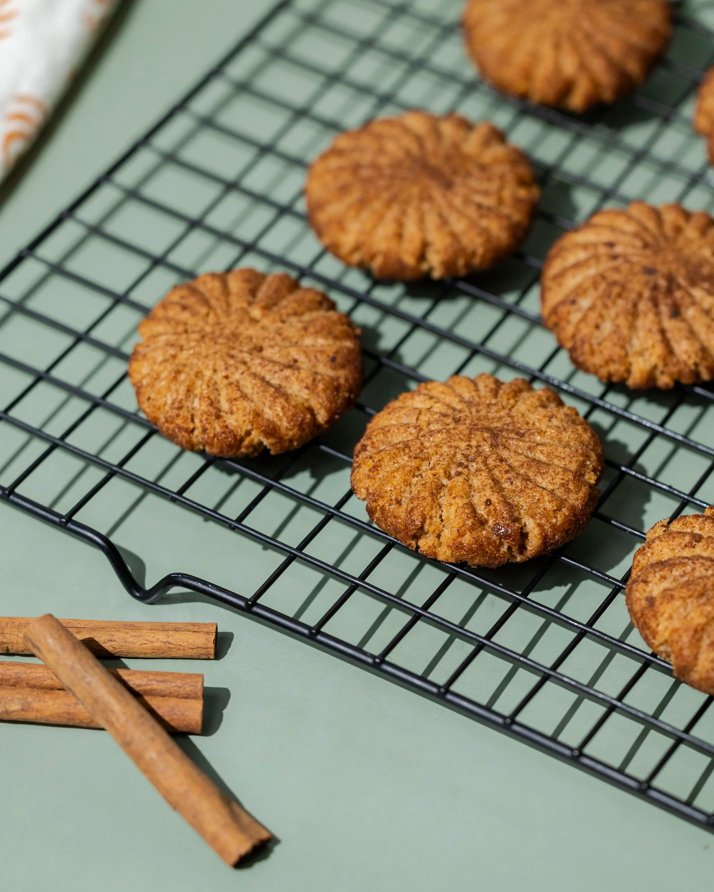 Close-up of several freshly baked cookies cooling on a black wire rack, with cinnamon sticks and a piece of wooden furniture nearby on a green surface.