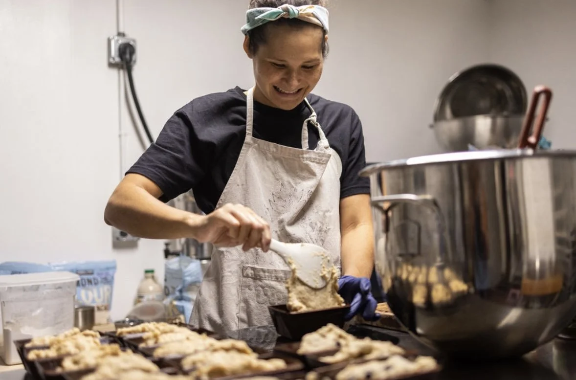 A woman wearing a headscarf, black t-shirt, and apron prepares baked goods in a kitchen, smiling as she scoops dough onto baking sheets.