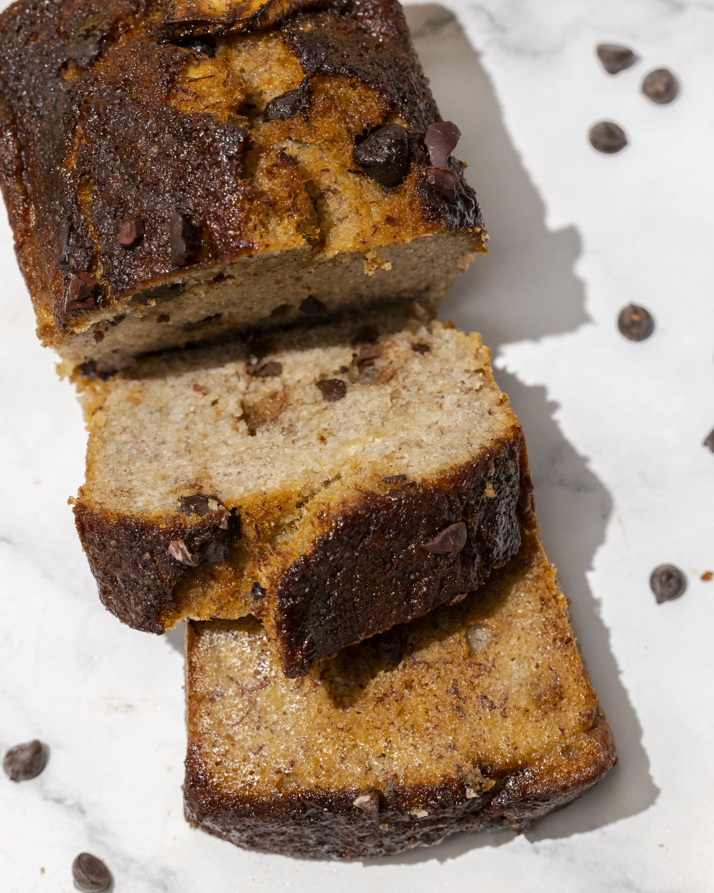 Close-up view of a stack of assorted banana bread slices topped with chocolate chips, on a white surface with scattered chocolate chips around.