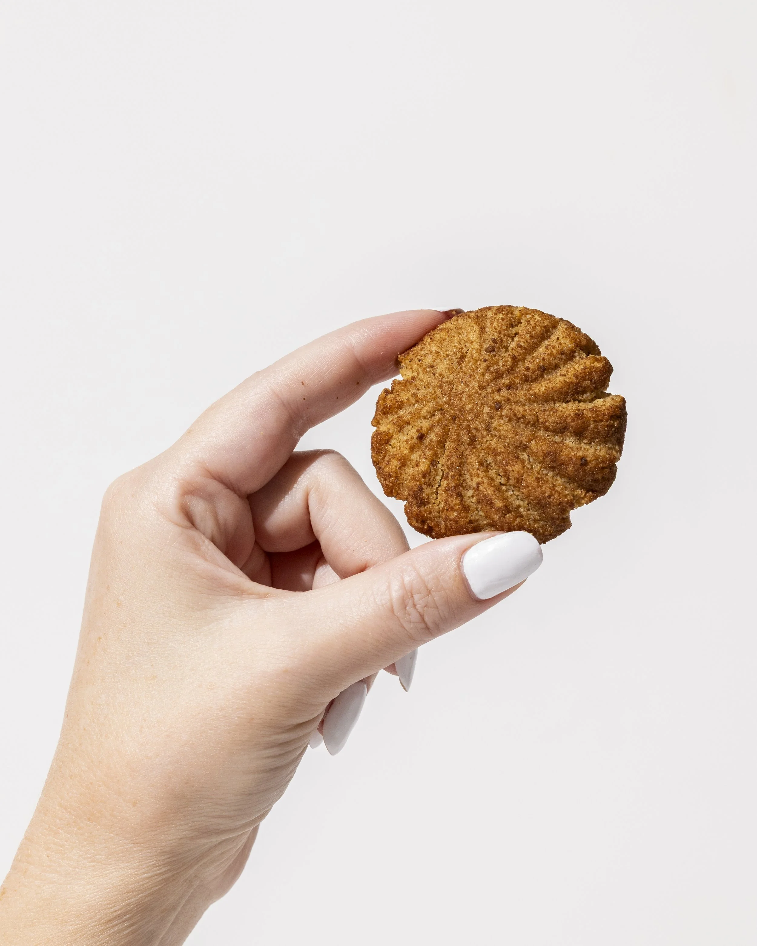 A hand with white painted nails holding a round, crispy, breaded cookie or biscuit against a plain white background.