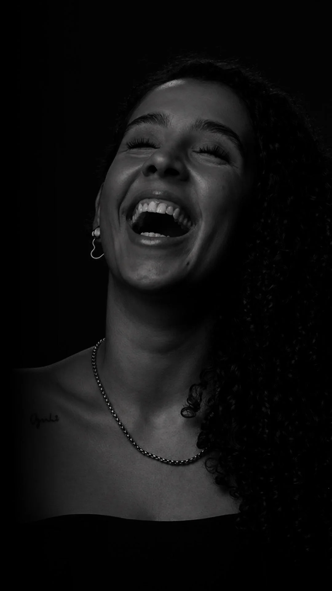 Black and white photo of a woman with curly hair, laughing with her eyes closed, showing her teeth, wearing earrings, necklace, and a strapless top.