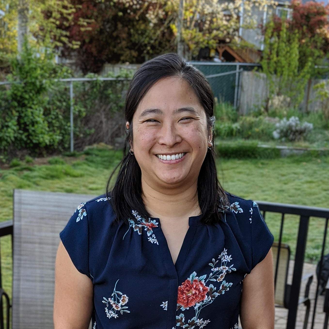 A woman smiling outdoors in a backyard with a fence, trees, and grass in the background.