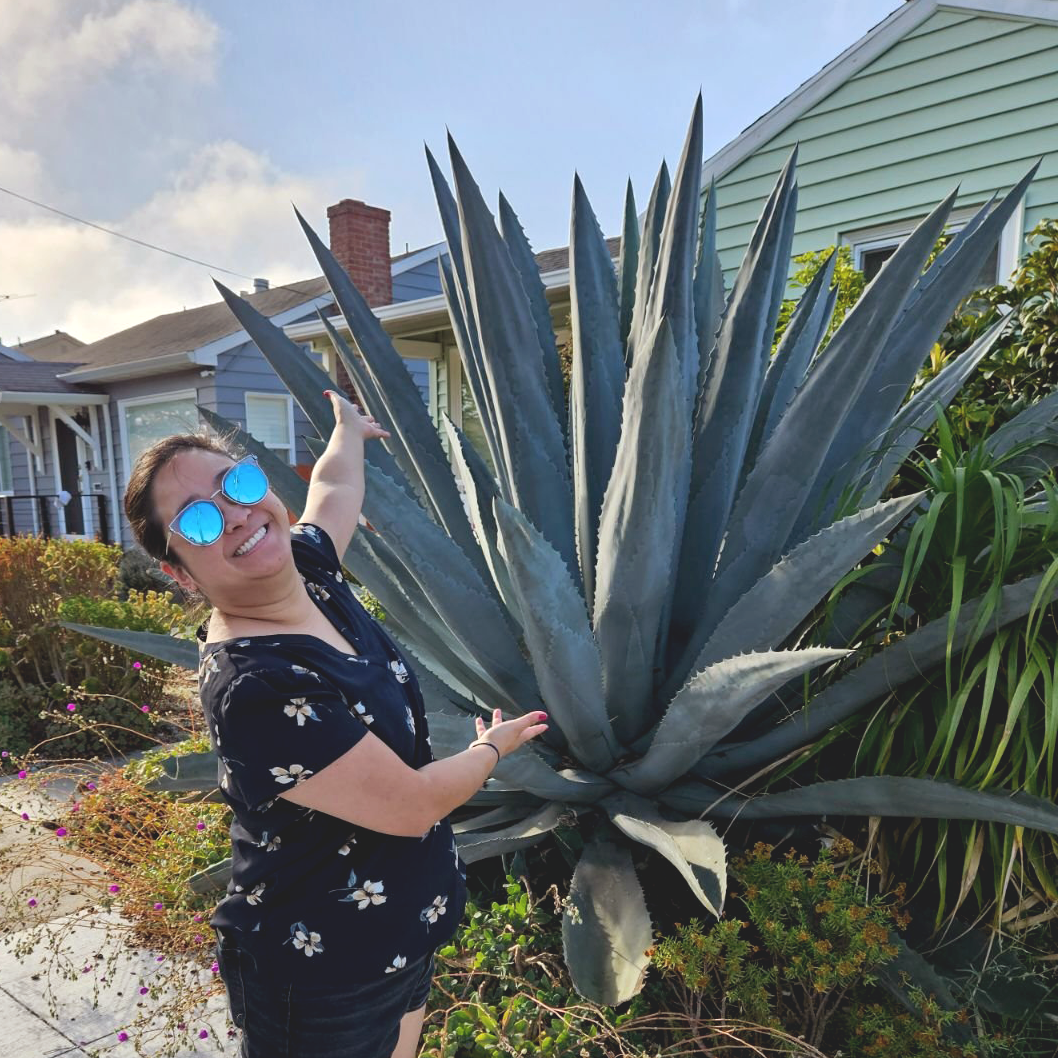 A woman wearing sunglasses and a floral shirt smiling and pointing at a large agave plant outside of a house.