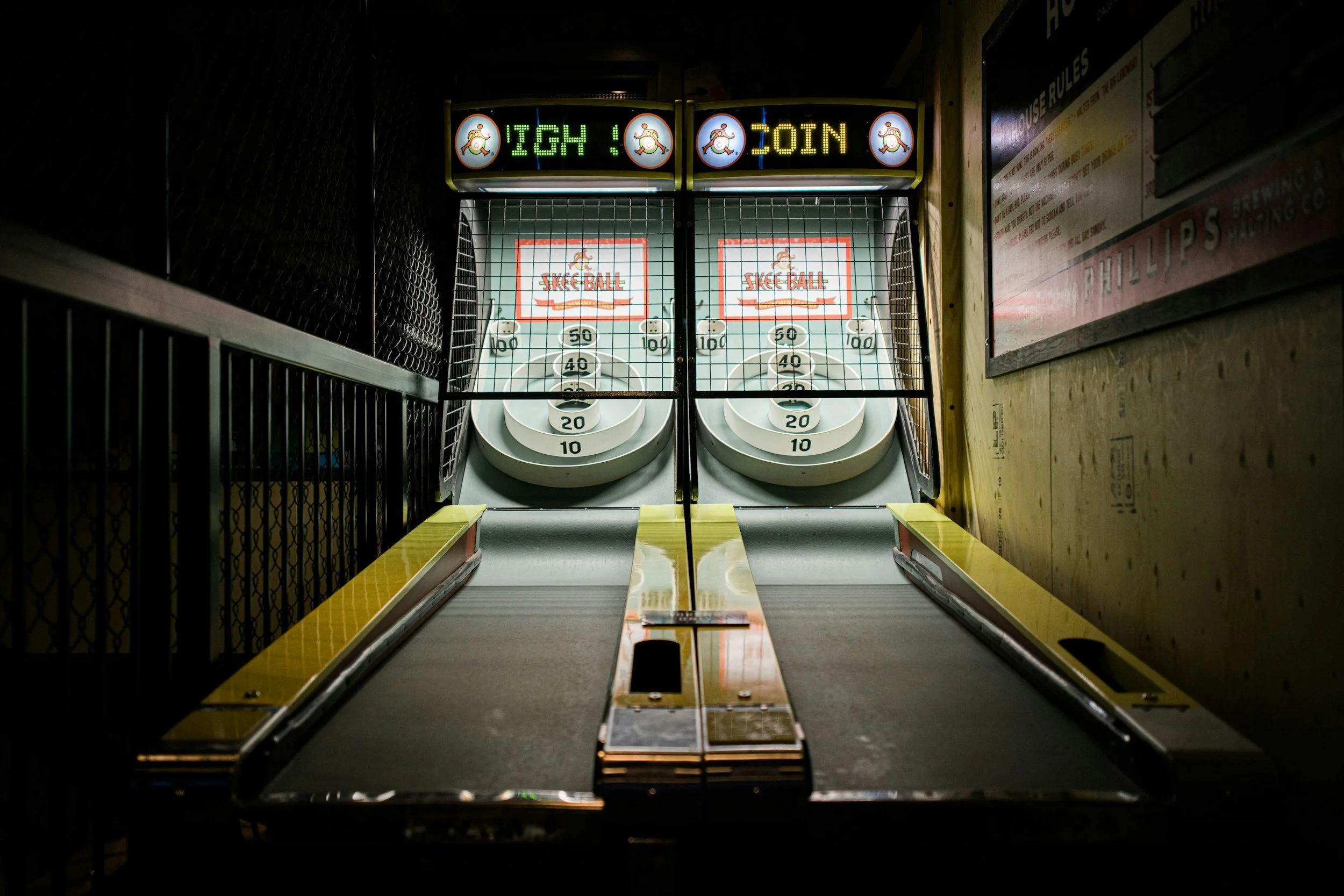 Indoor skee ball alley with digital scoreboard overhead reading 'High Join' with images of a squirrel. Two skee ball lanes with numbered circular target areas and white balls, surrounded by a yellow safety border.