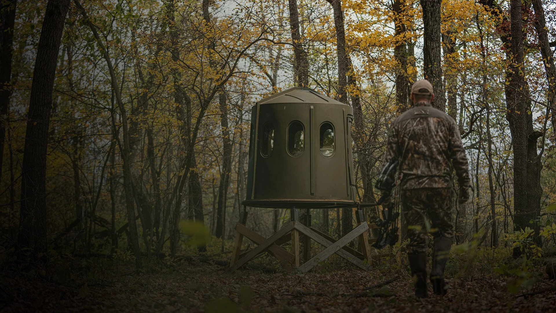 A person in camouflage clothing walking through a forest towards a hunting blind elevated on a wooden platform during autumn.