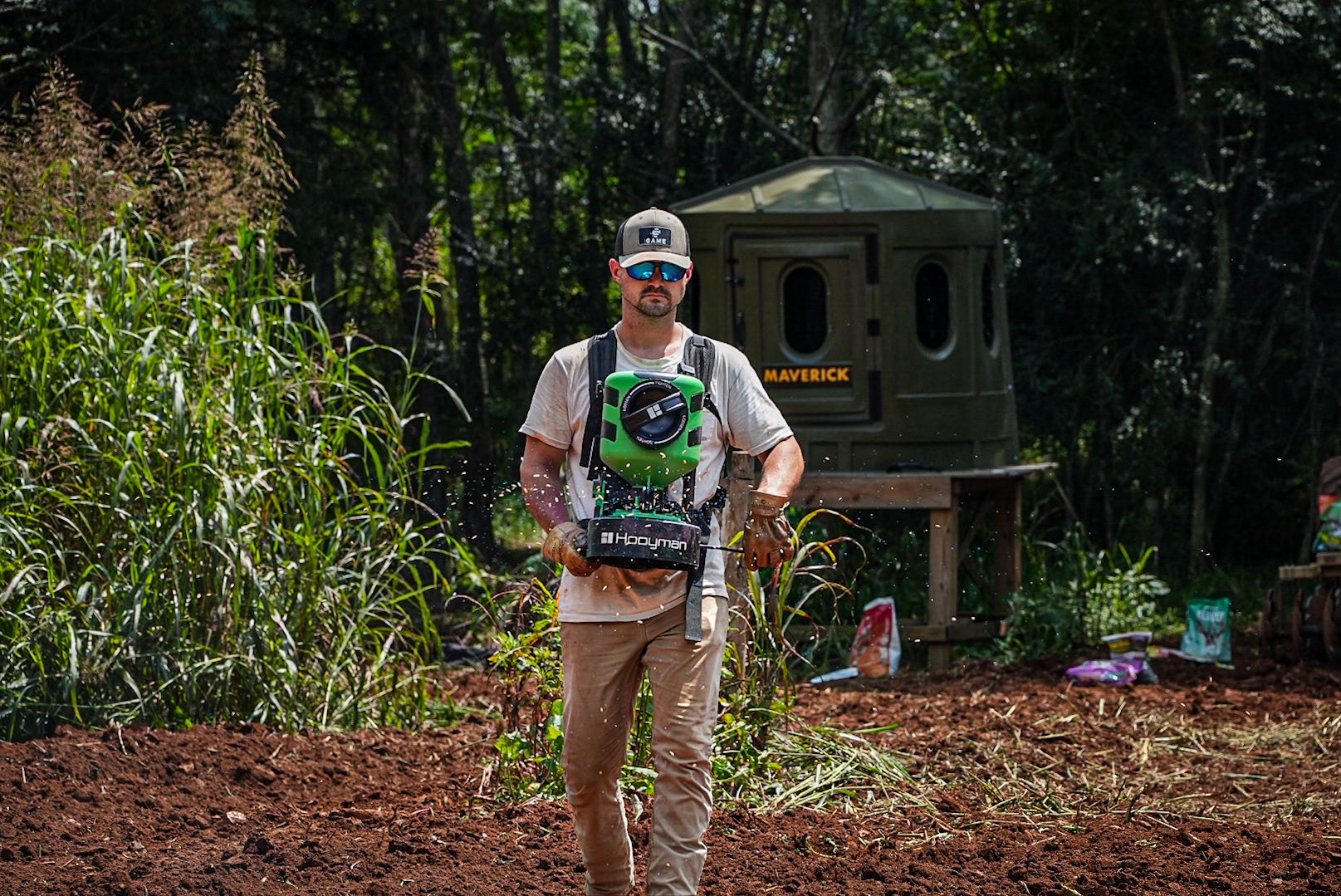 A man with sunglasses and gloves is spraying pesticides in a garden with a backpack sprayer, surrounded by greenery and plants.