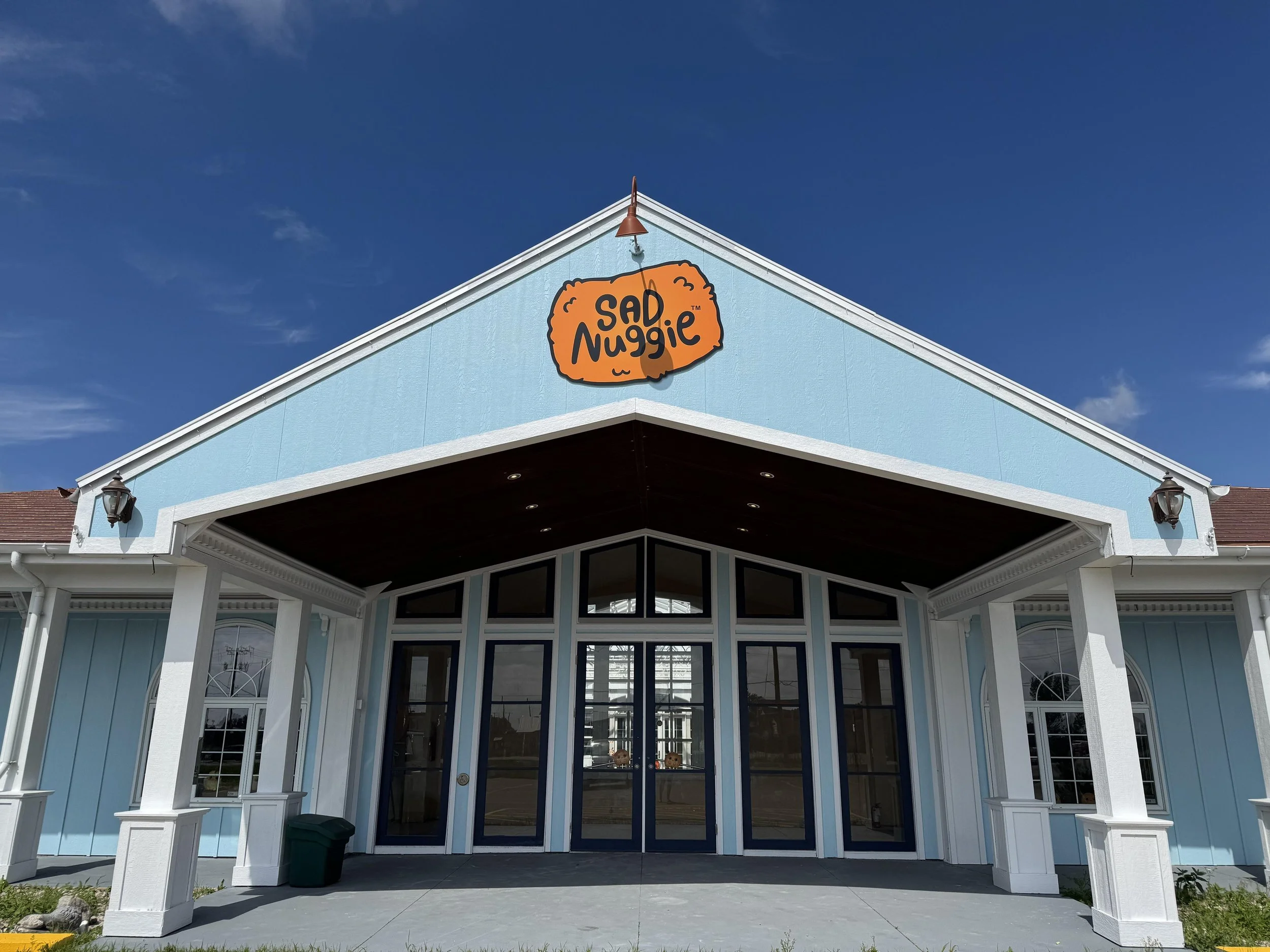 Front view of a building with a blue exterior, white trim, and a sign that reads 'Sad Nugget' on the gable peak. The building has large glass doors and windows, with a covered entrance. The sky is clear and blue.