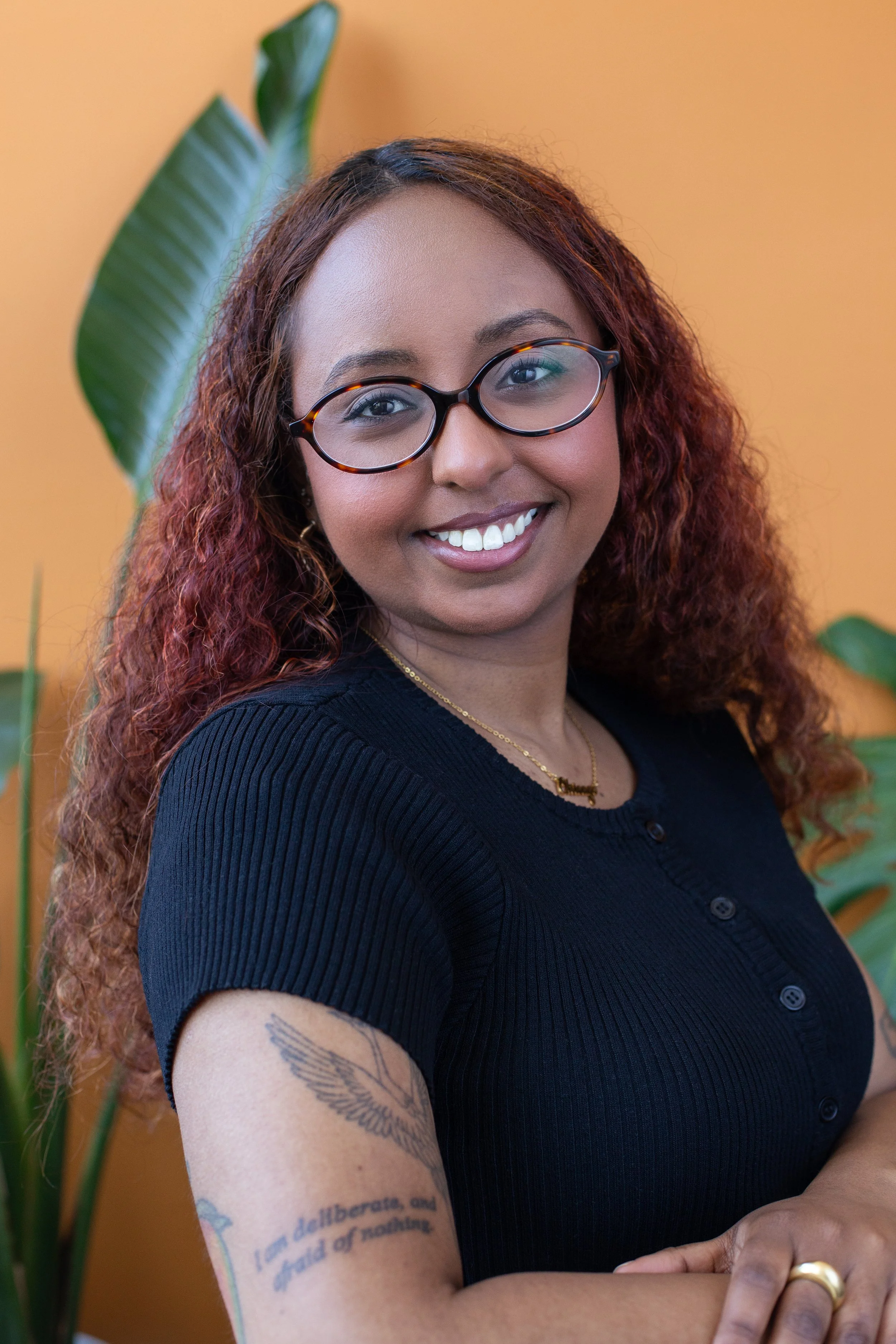 Black woman with glasses posed with orange-colored background and green leaves