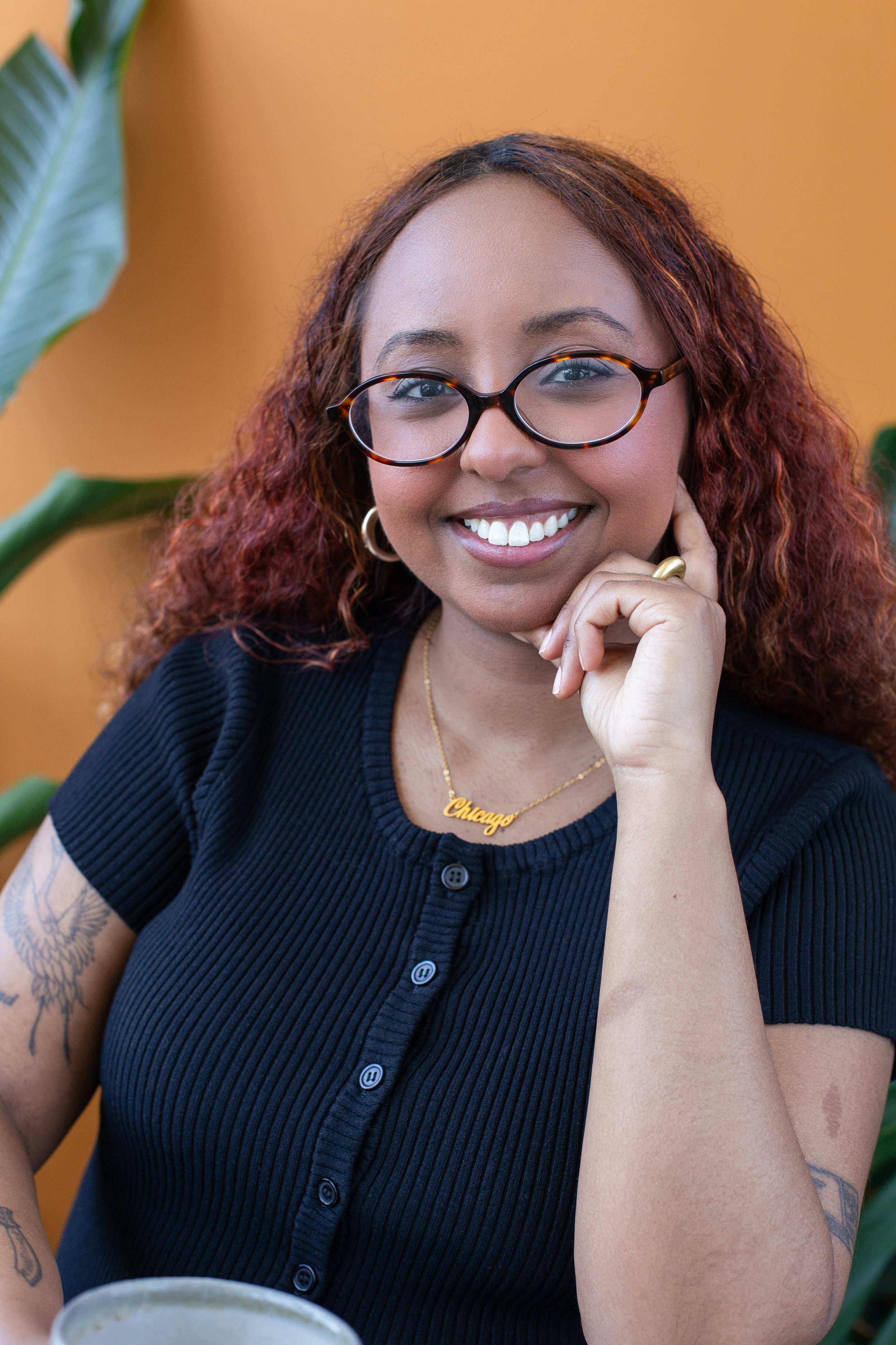 Black woman posed with gold jewelry, orange-colored backgroud, and oval-shaped glasses