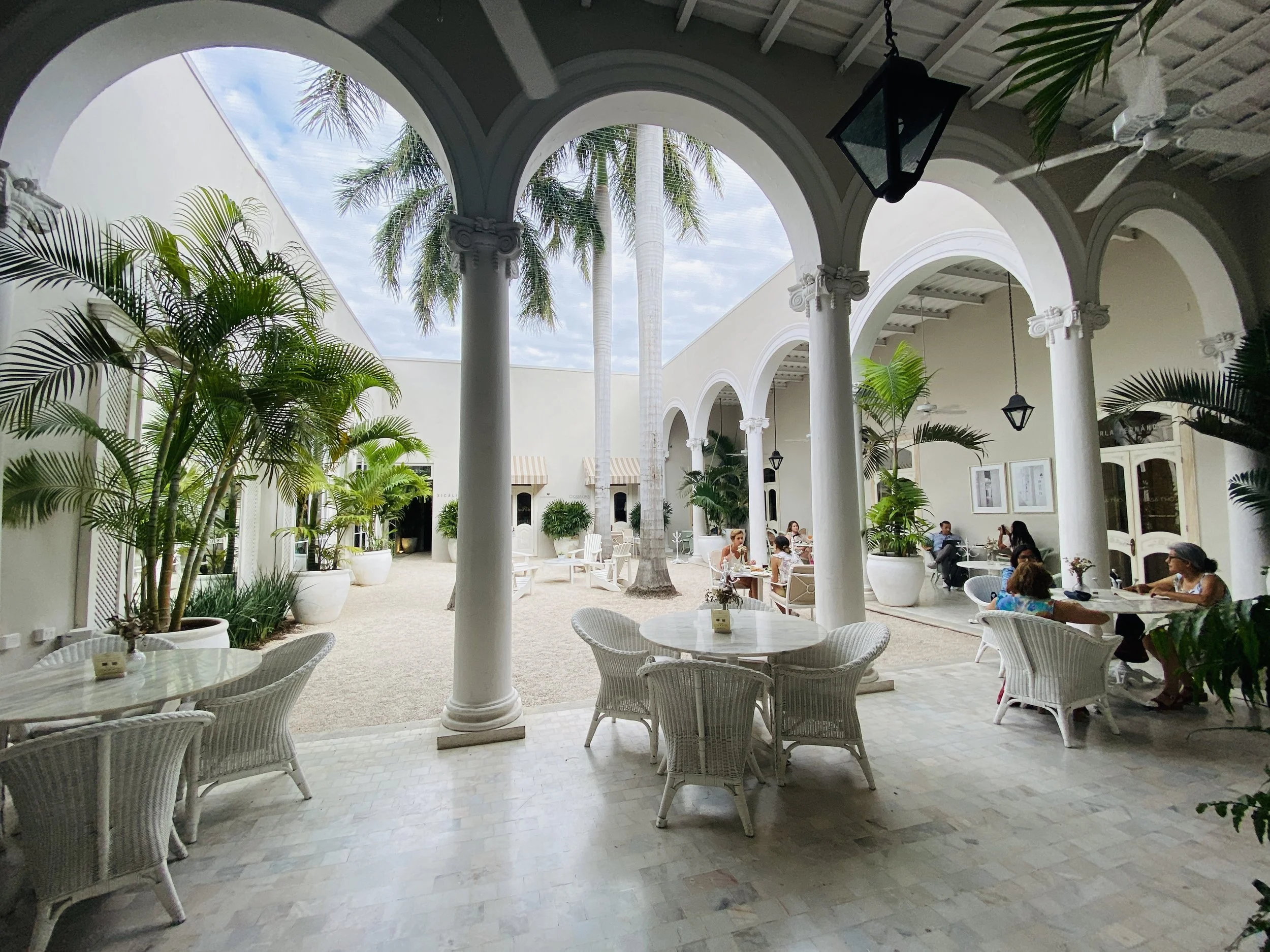 People sitting at cafe tables in the white arched courtyard of Casa T'HŌ Concept House on Paseo de Montejo in Mérida, Yucatán, Mexico