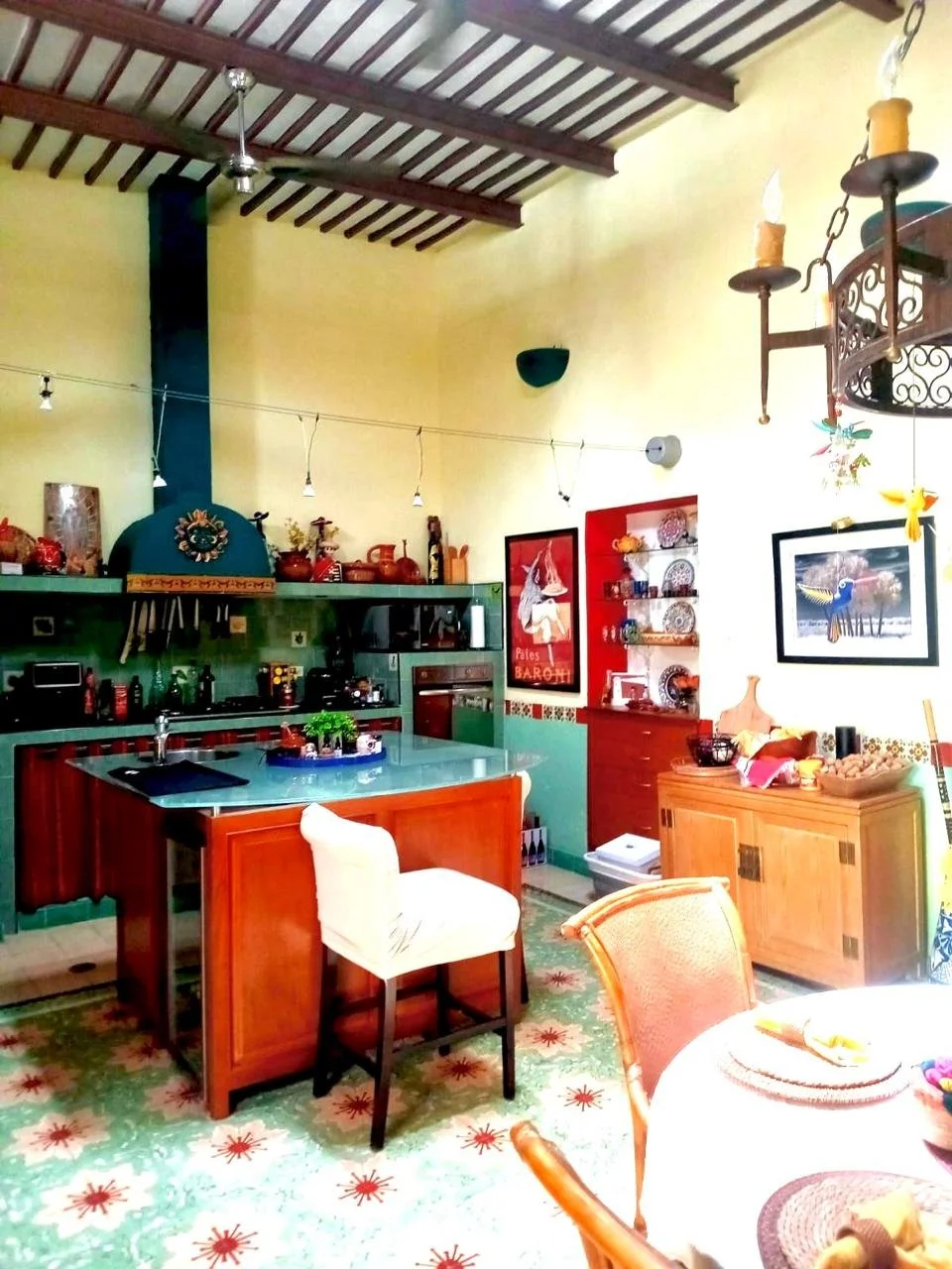 A colorful kitchen with yellow walls, green cabinets, a wooden kitchen island, and a dining area with chairs and a table. Decor includes framed artwork, hanging candles, and decorative items on shelves.