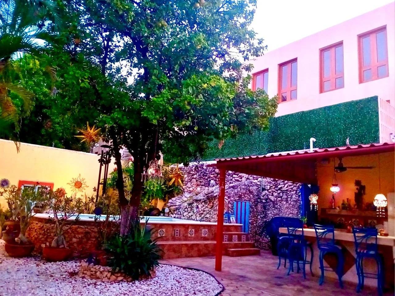 A vibrant outdoor patio with a large tree, potted plants, a covered seating area with blue chairs, a stone wall, and a multi-story building with pink window frames in the background.