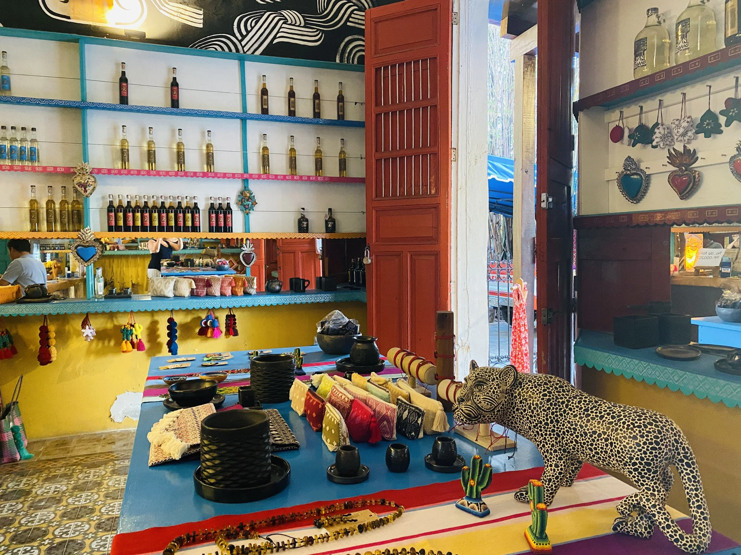 Brightly decorated Mexican Posheria boutique shop on the Paseo de Montejo with colorful tables displaying handcrafted textiles, jewelry, a spotted jaguar sculpture, and shelves lined with artisanal bottles.