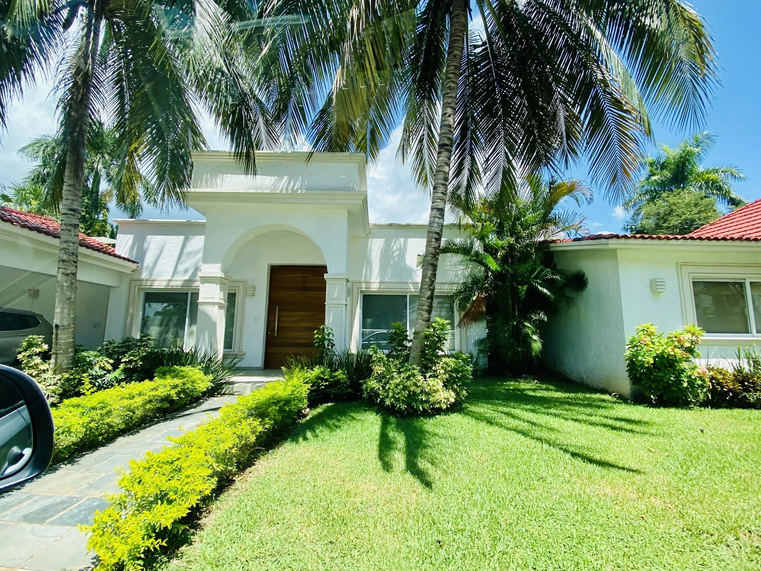Bright white single-story modern villa in La Ceiba golf community with a wooden front door, arched entryway, lush green lawn, and towering  palm trees under a blue sky.