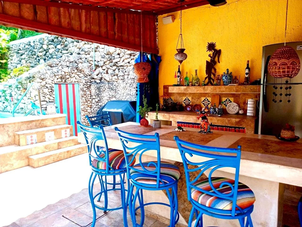 Colorful outdoor kitchen with a yellow wall, a bar counter with decorative items, and bright blue chairs with striped cushions around a mosaic-topped table. Stone steps lead to a rocky backdrop with a striped door or screen.