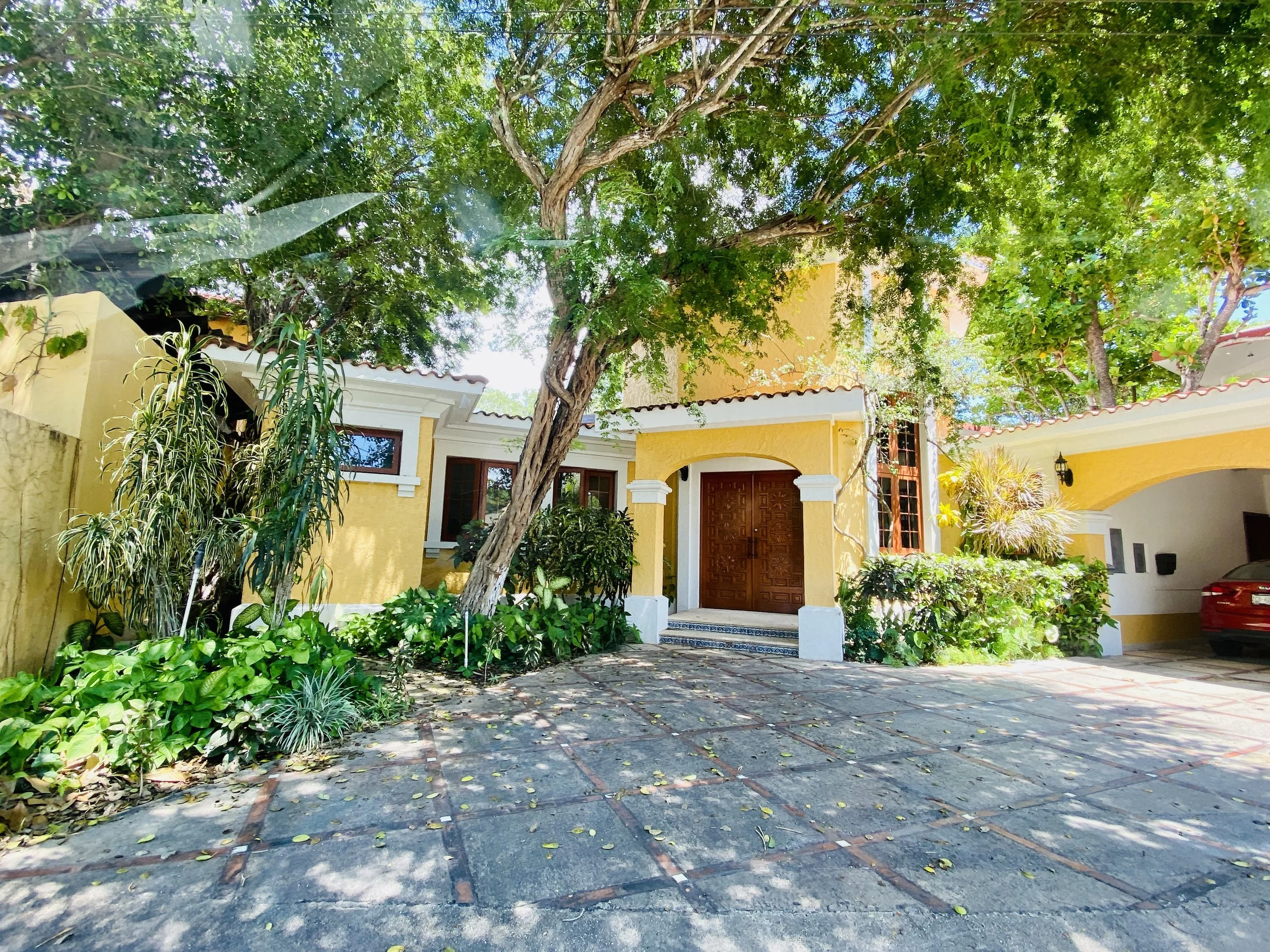 Bright yellow colonial-style home in La Ceiba golf community with ornate double wooden front doors, lush tropical trees, and a spacious paved driveway.