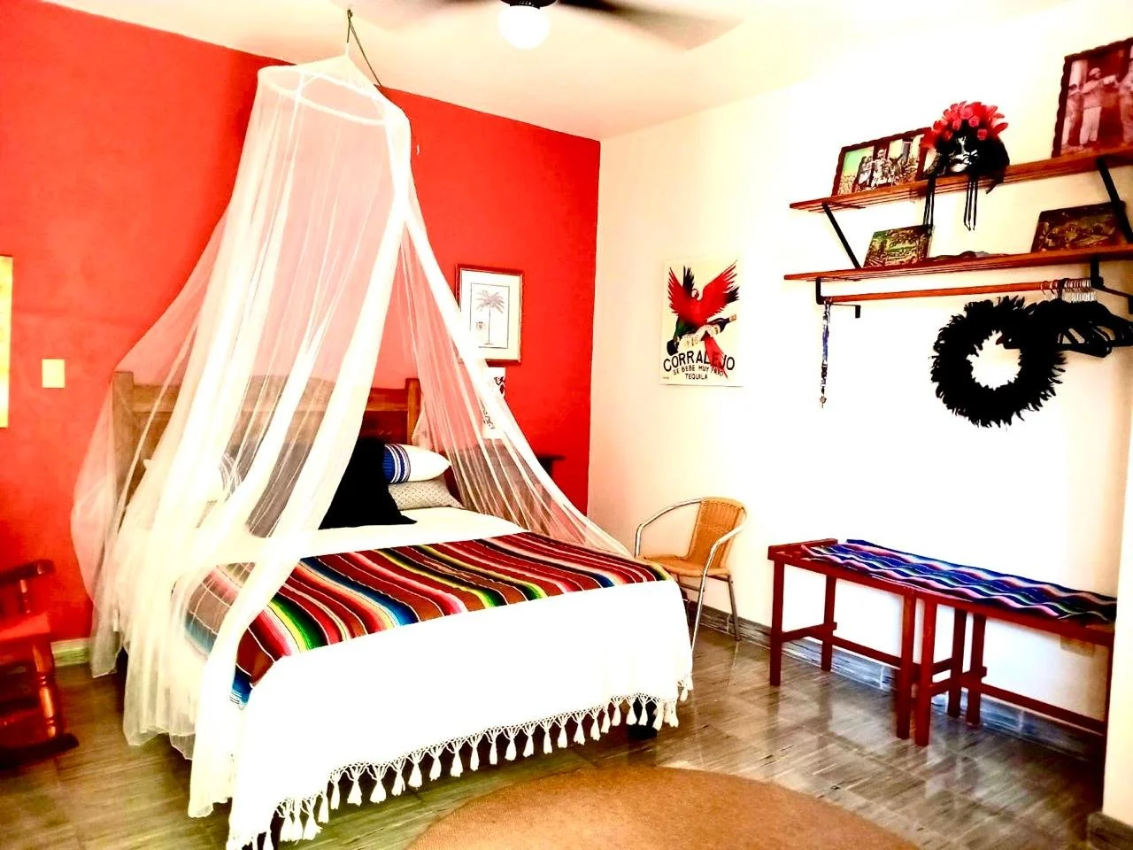 A bedroom with a red accent wall, a bed with Mexican-style blanket, a mosquito net canopy, a wicker chair, a wooden table with a colorful tablecloth, and wall decorations including framed pictures, a poster, and a black circular wreath.