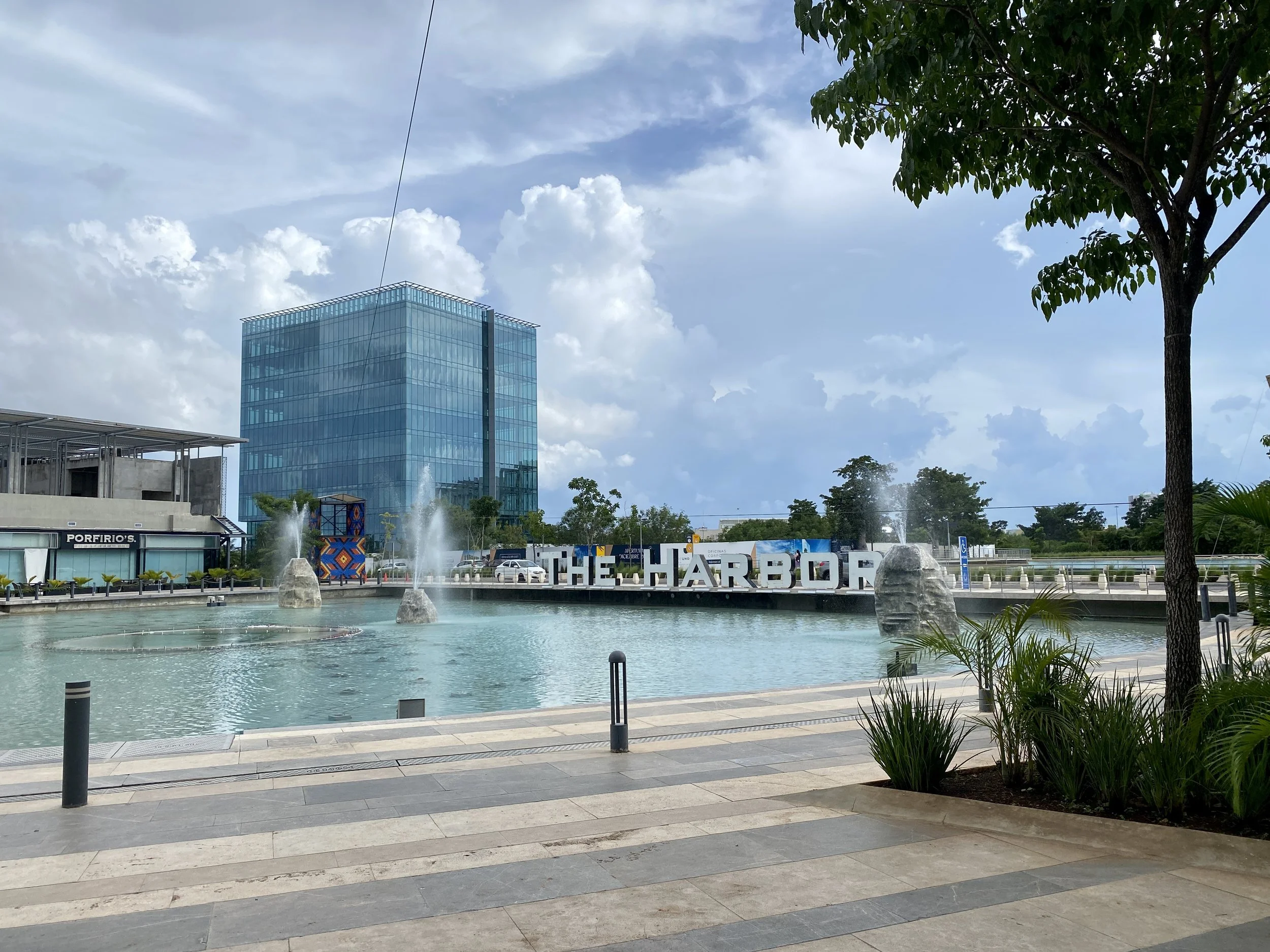 Large outdoor decorative lake with water fountains in front of "THE HARBOR" sign and a modern blue glass office tower at The Harbor Mérida shopping mall.