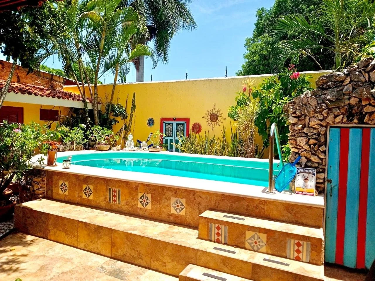A backyard swimming pool surrounded by tropical plants and colorful decorations. There is a yellow wall with sun and flower art, and a door with a red and blue border. The pool has a tiled border and a small set of steps leading into it. On the right, there is a wooden gate with red and teal stripes, a pool cleaning net, and a warning sign with a dog image.