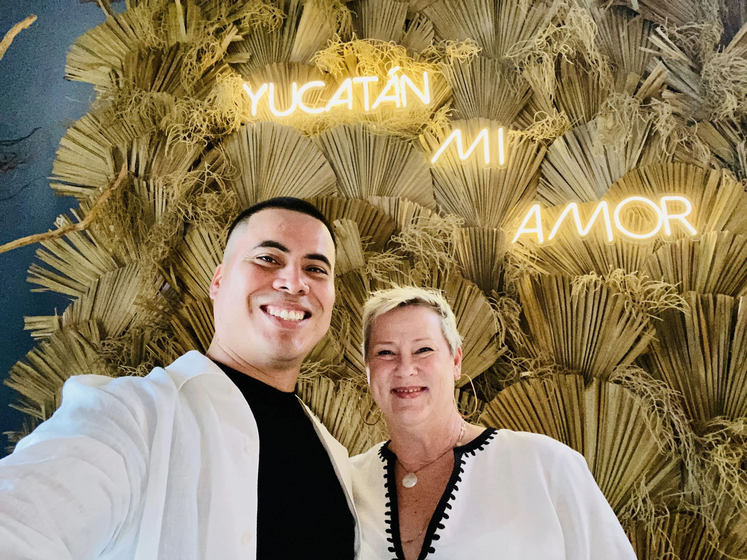 Angel Rodriguez and Amy Jones, Co-Founders of Life in Mérida™ standing in front of a wall of fans and a Yucatán Mi Amor neon sign