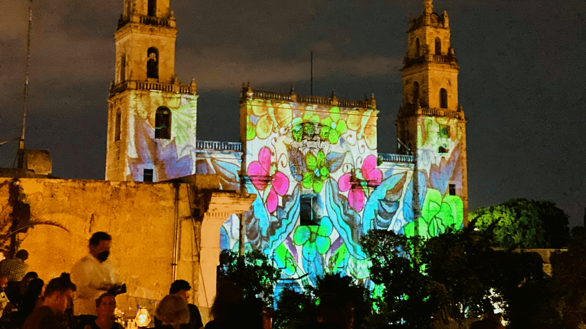 The Mérida Cathedral at night illuminated by a colorful, artistic video mapping light show, viewed from a rooftop with people in the foreground.