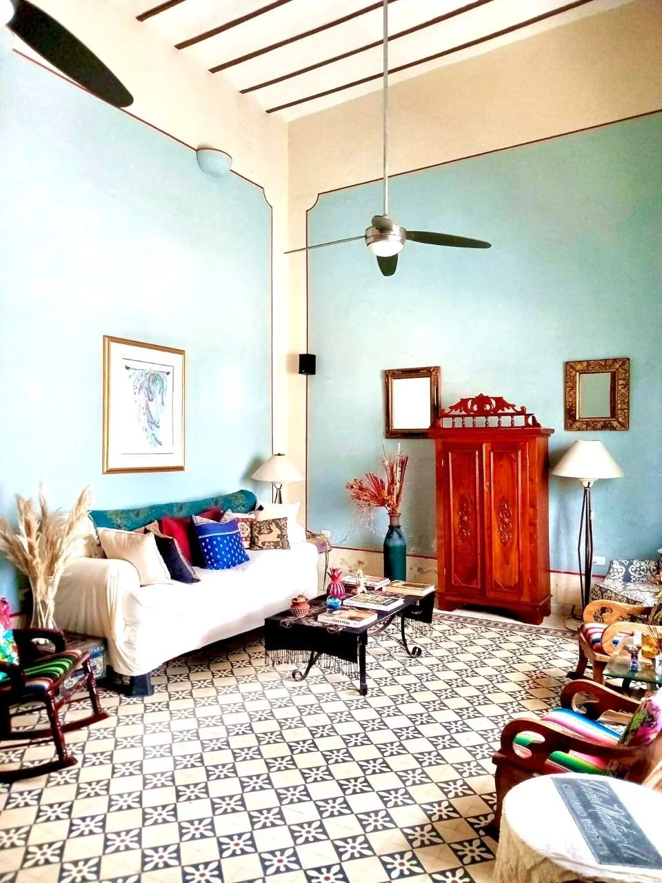Living room with high ceiling and ceiling fan, blue walls, white sofa with multicolored cushions, wooden furniture, patterned floor, and various decorative items.