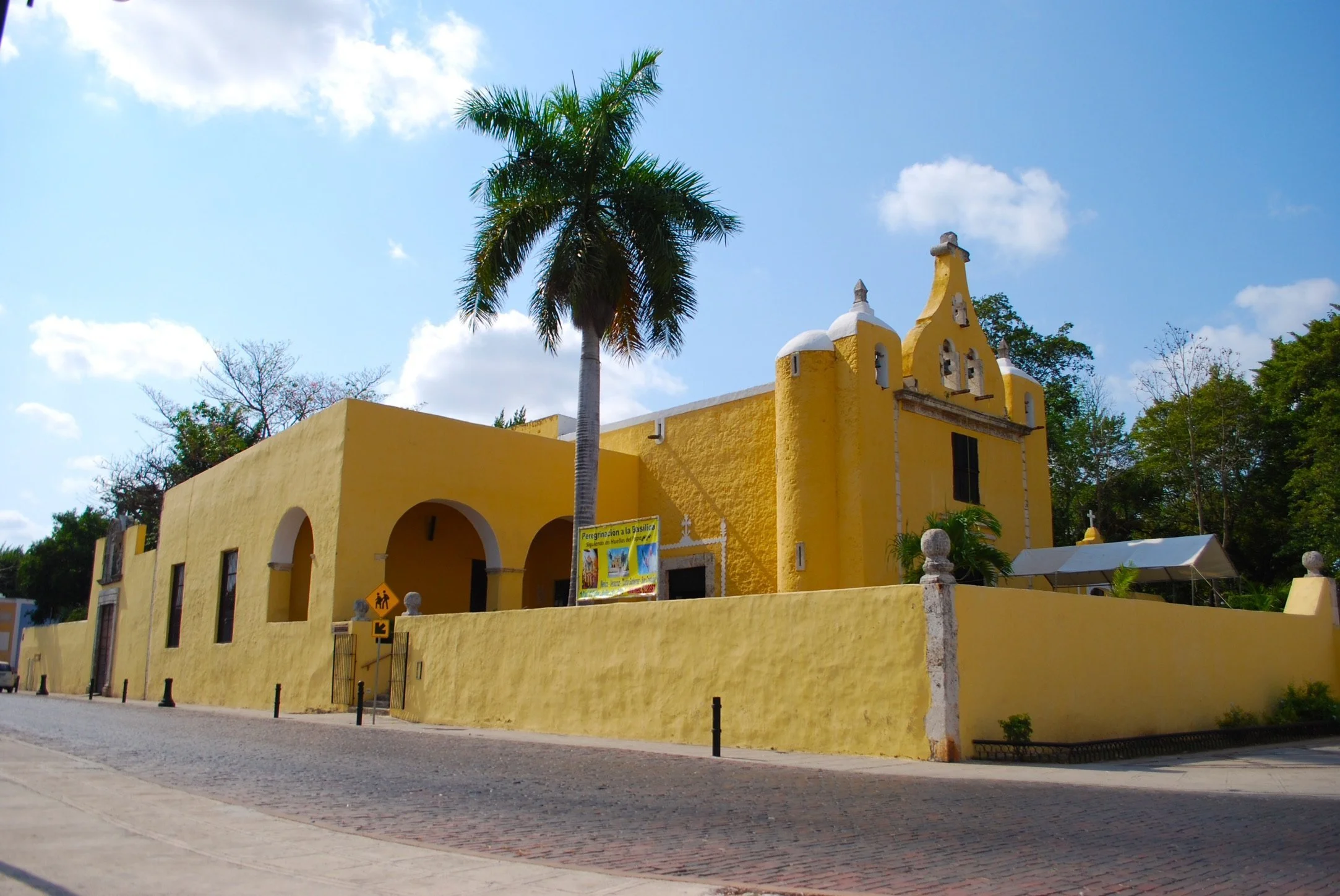 Exterior view of the bright yellow Ermita de Isabel Church in Mérida, with a tall palm tree standing beside its stone wall on a sunny day.