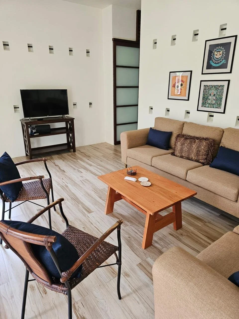 Living room with beige sofa, black pillows, a wooden coffee table, and a TV on a stand. Walls decorated with framed artwork, wood and glass partition, and wooden flooring.