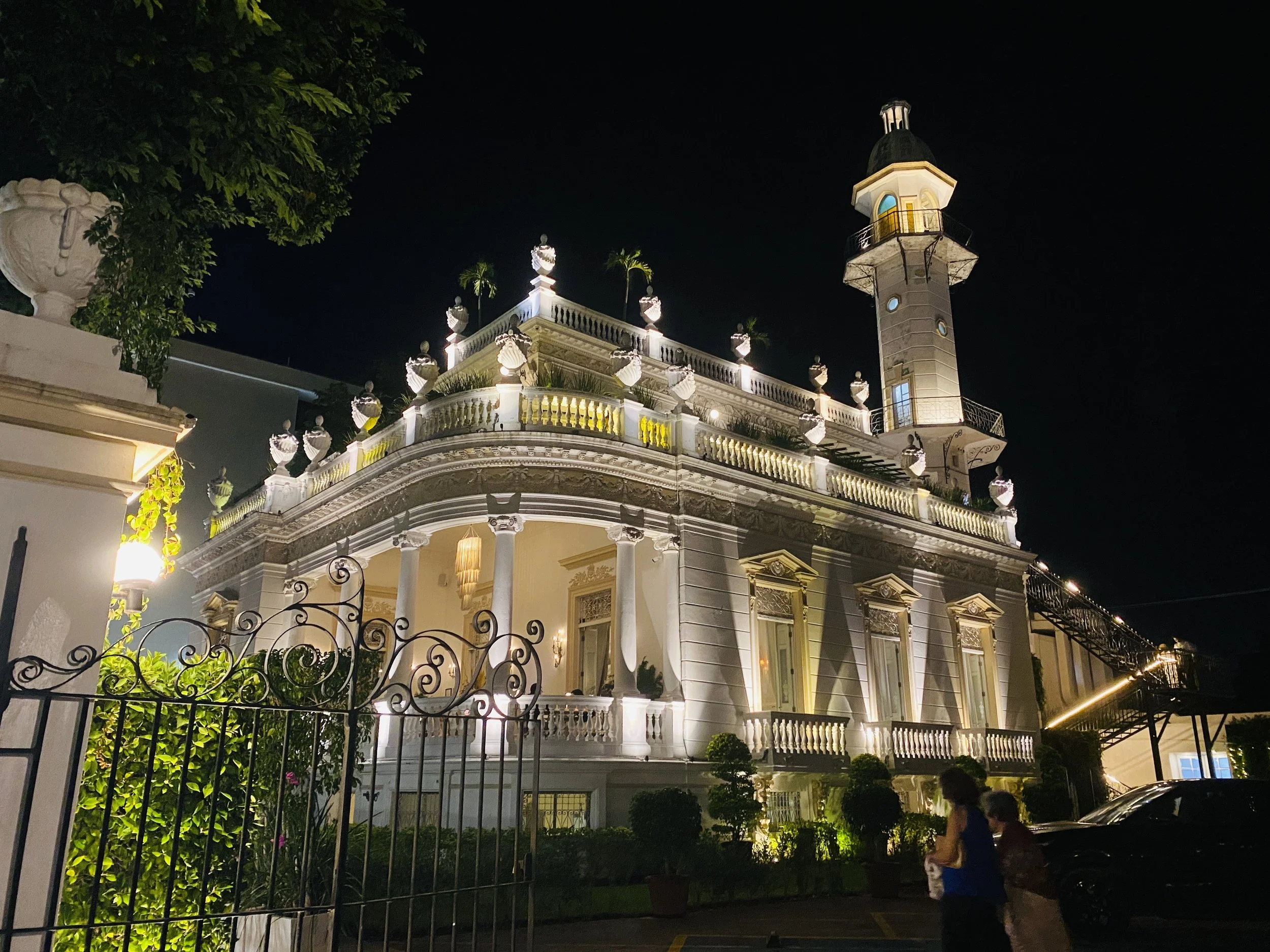 Exterior view of El Minaret mansion on the Paseo de Montejo  at night, illuminated with golden lights, featuring a distinctive minaret tower and classical columns.