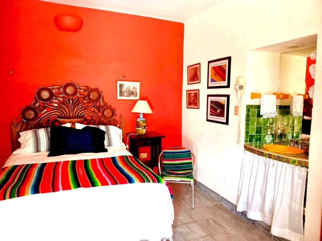 A bedroom with a red accent wall, ornate wooden headboard, striped bedding, and black pillows. Beside the bed is a small wooden nightstand with a lamp, and to the right, framed artwork on a white wall. There's a small colorful striped chair and a sink with green tiles and a white curtain below. The room has a warm, vibrant decor style.