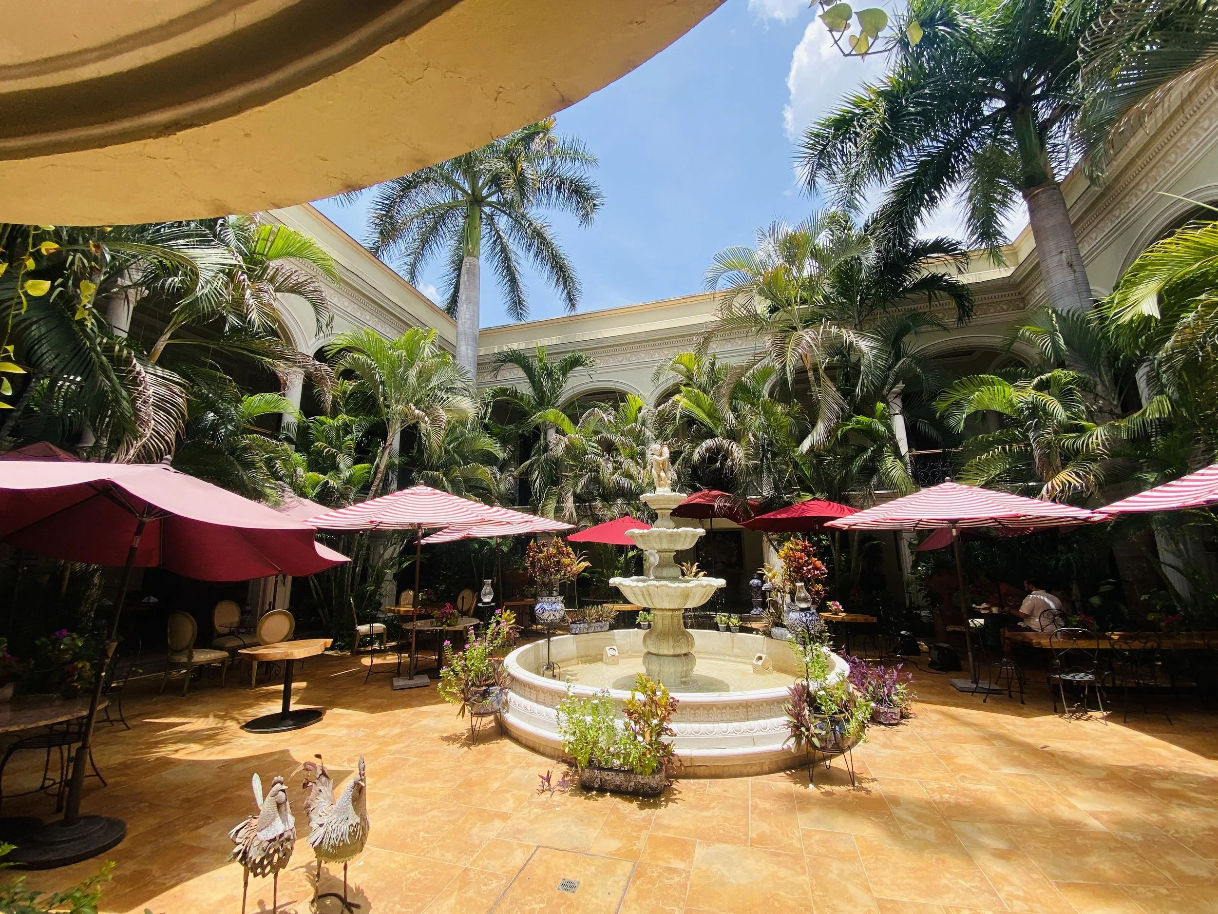 Sunlit inner courtyard of a colonial boutique hotel featuring a central tiered stone fountain, lush palm trees, and patio seating under red umbrellas.
