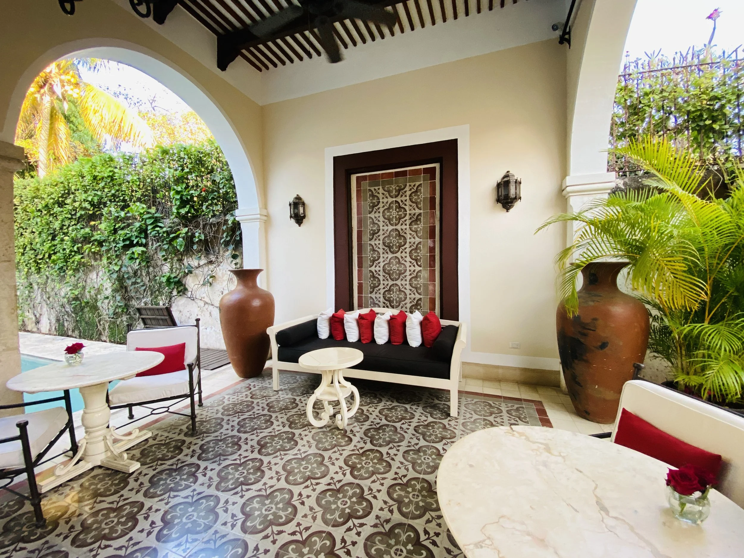 Covered outdoor terrace featuring patterned cement tile flooring, arched doorways, a sofa with red and white pillows, small bistro tables, and large clay urns near a pool.