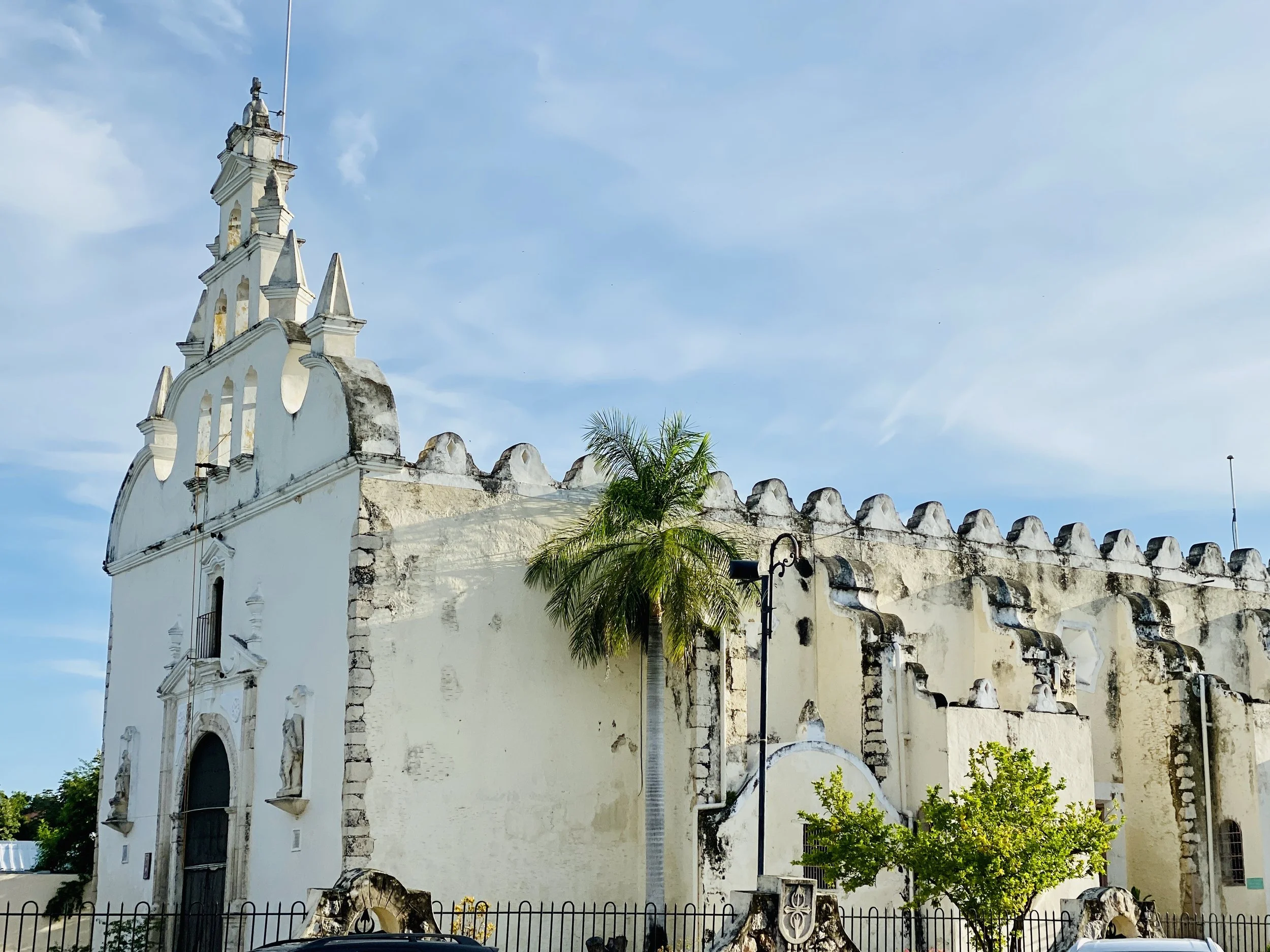 Exterior view of the historic white Santiago Church (Iglesia de Santiago Apóstol) in Mérida, featuring a multi-tiered belfry wall and palm trees under a bright sky.