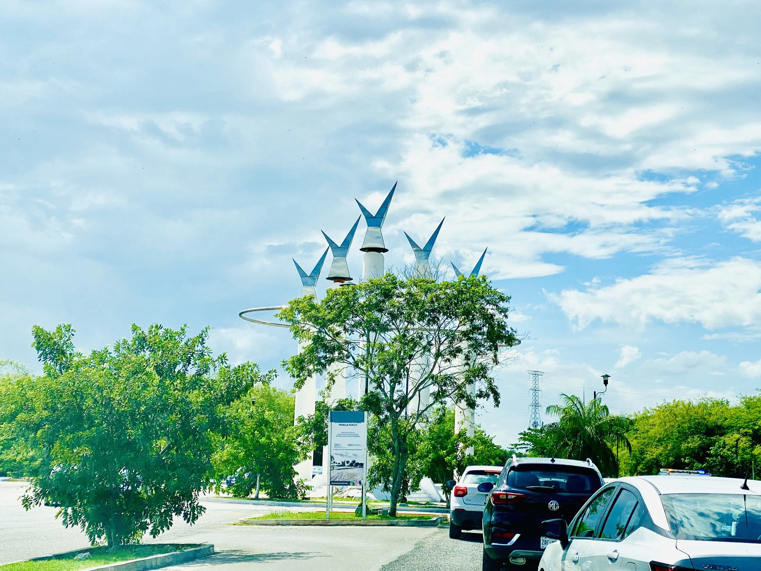 A modern outdoor sculpture with five white pillars topped with metallic finials, surrounded by trees and traffic at a roundabout under a bright sky.