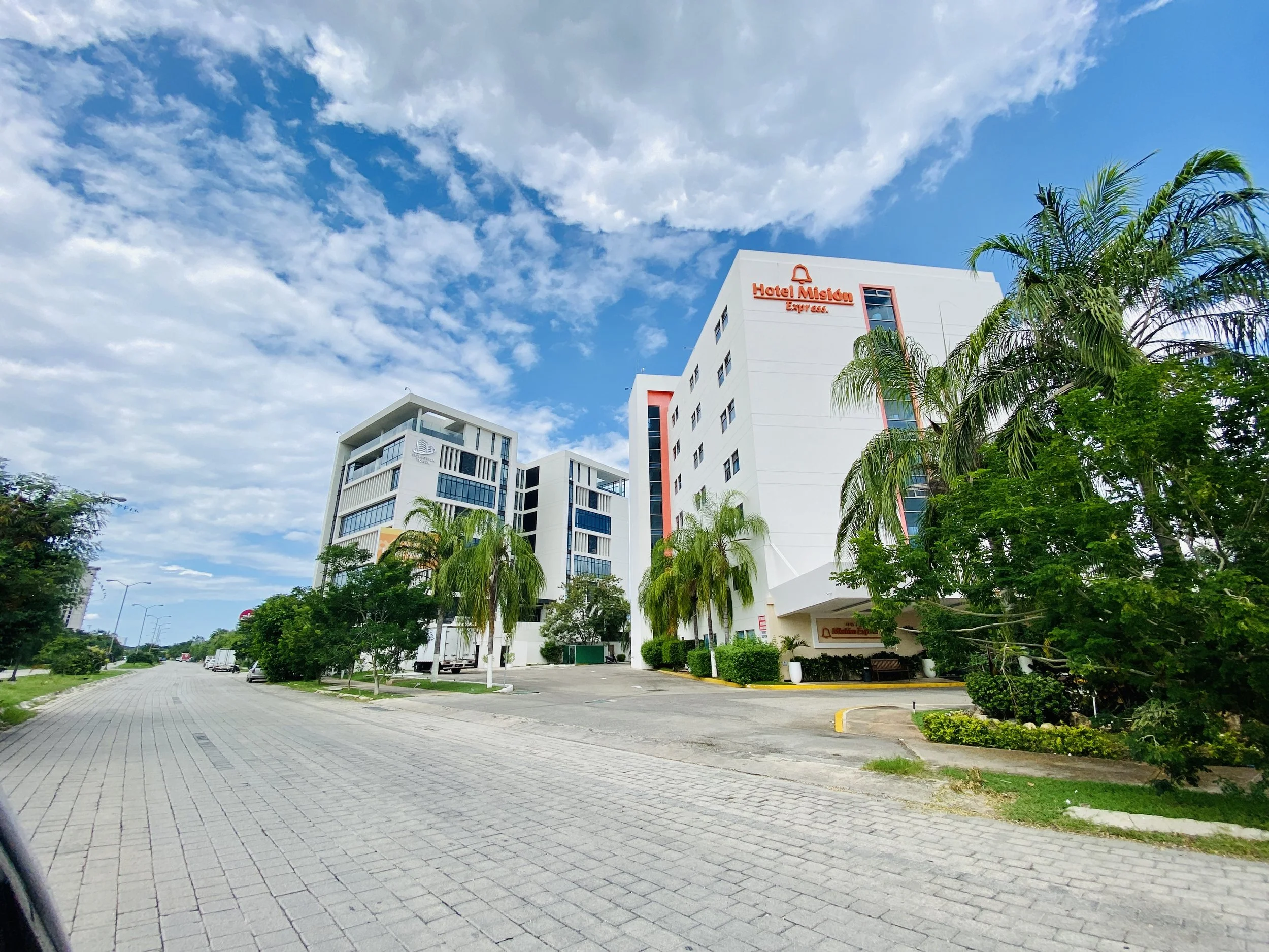 Exterior view of Hotel Misión Express in the Altabrisa neighborhood, a multi-story modern white building with orange signage along a paved street lined with palm trees under a blue sky.