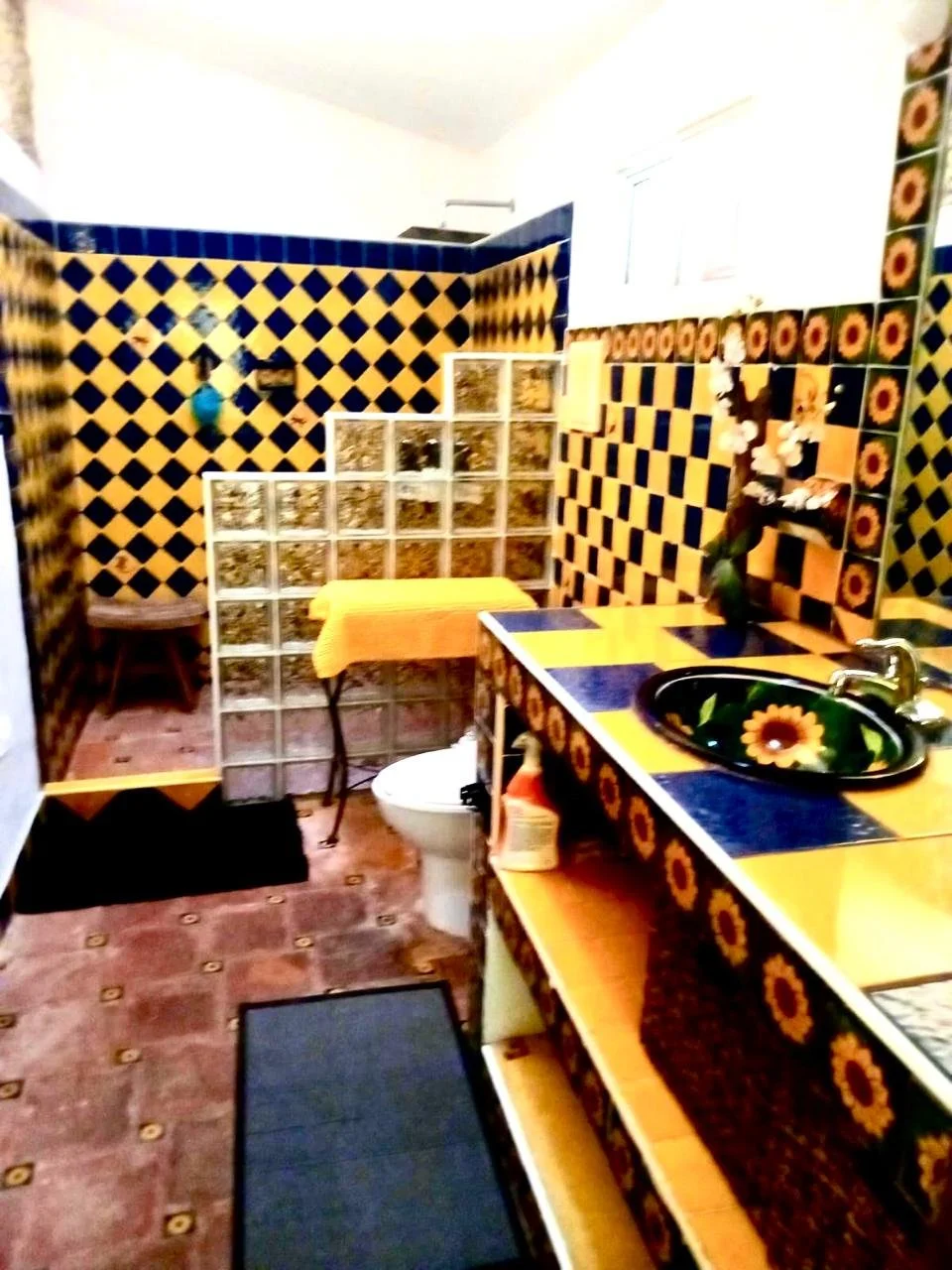 A bathroom with yellow and blue tiles, sunflower-themed decor, a glass block partition, and a black sink with sunflower design.