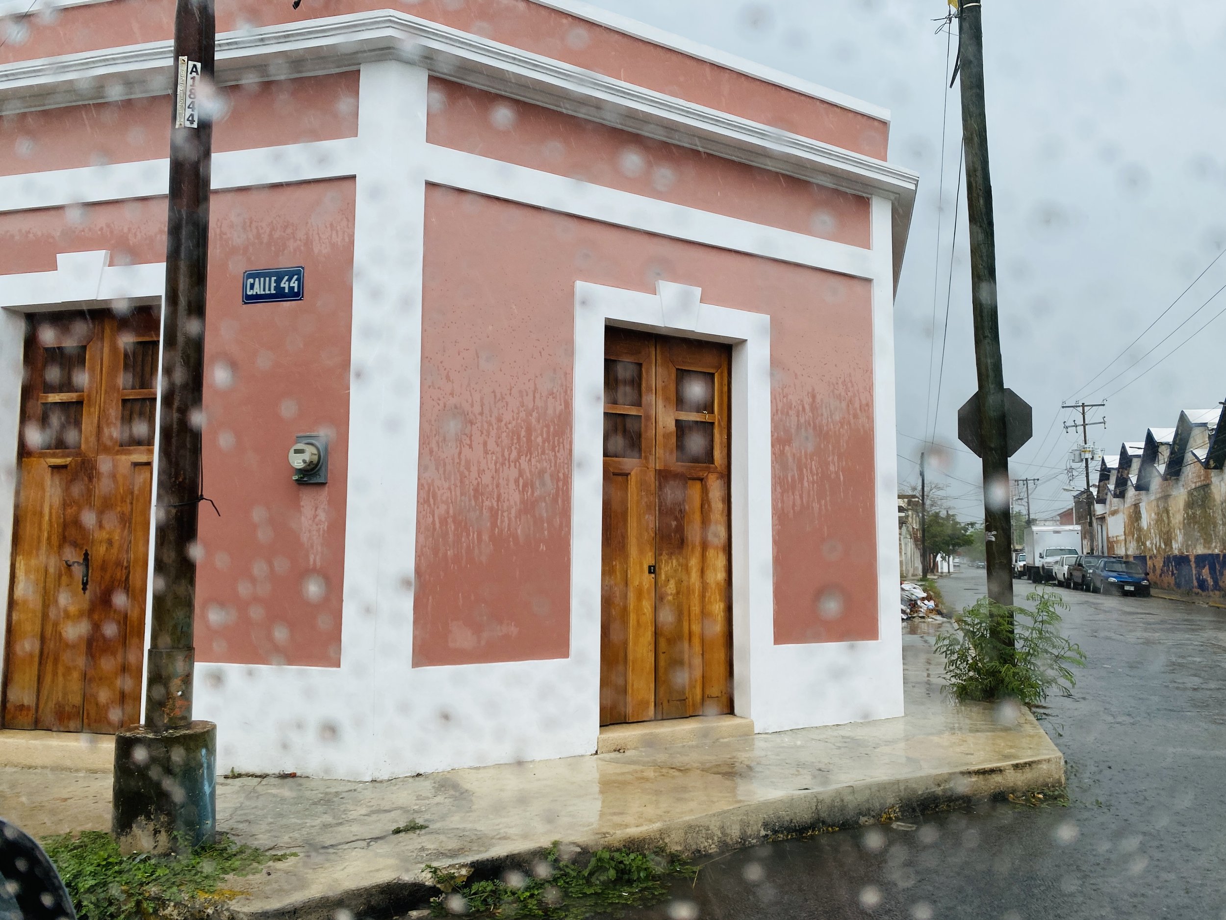 A pink building with white trim and wooden doors on a rainy day. There is a street sign that reads 'Calle 44' on a pole in front of it. Rain droplets are visible on the window through which the photo is taken. The street appears wet and there are some small plants at the base of the building.