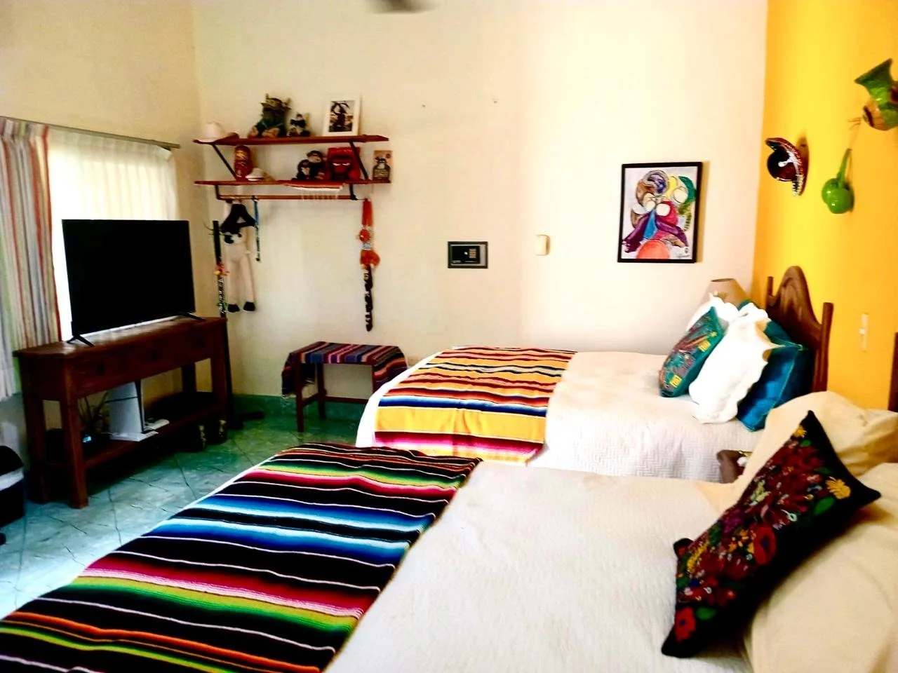 A hotel room with two double beds featuring colorful striped bed runners and pillows, a wooden TV stand with a flat screen TV, a wall-mounted shelf with decorative items, colorful masks on the wall, and a piece of framed artwork, with green-tiled flooring and a window with striped curtains.