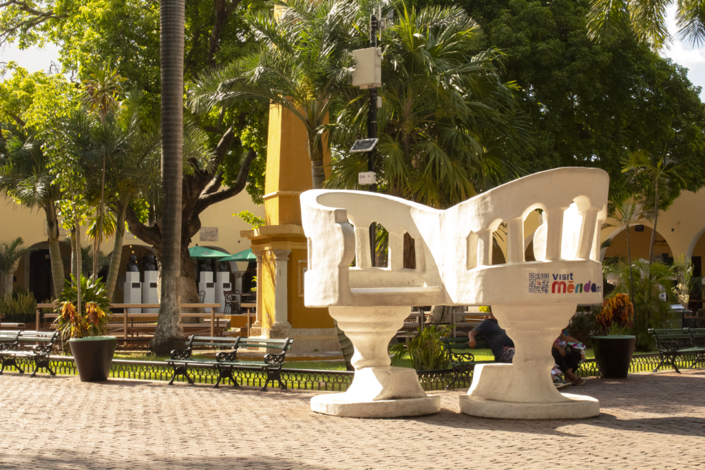 Giant white "kissing chair" sculpture (silla confidente) in Parque Santa Lucía, Mérida, set against a sunny park backdrop with palm trees.