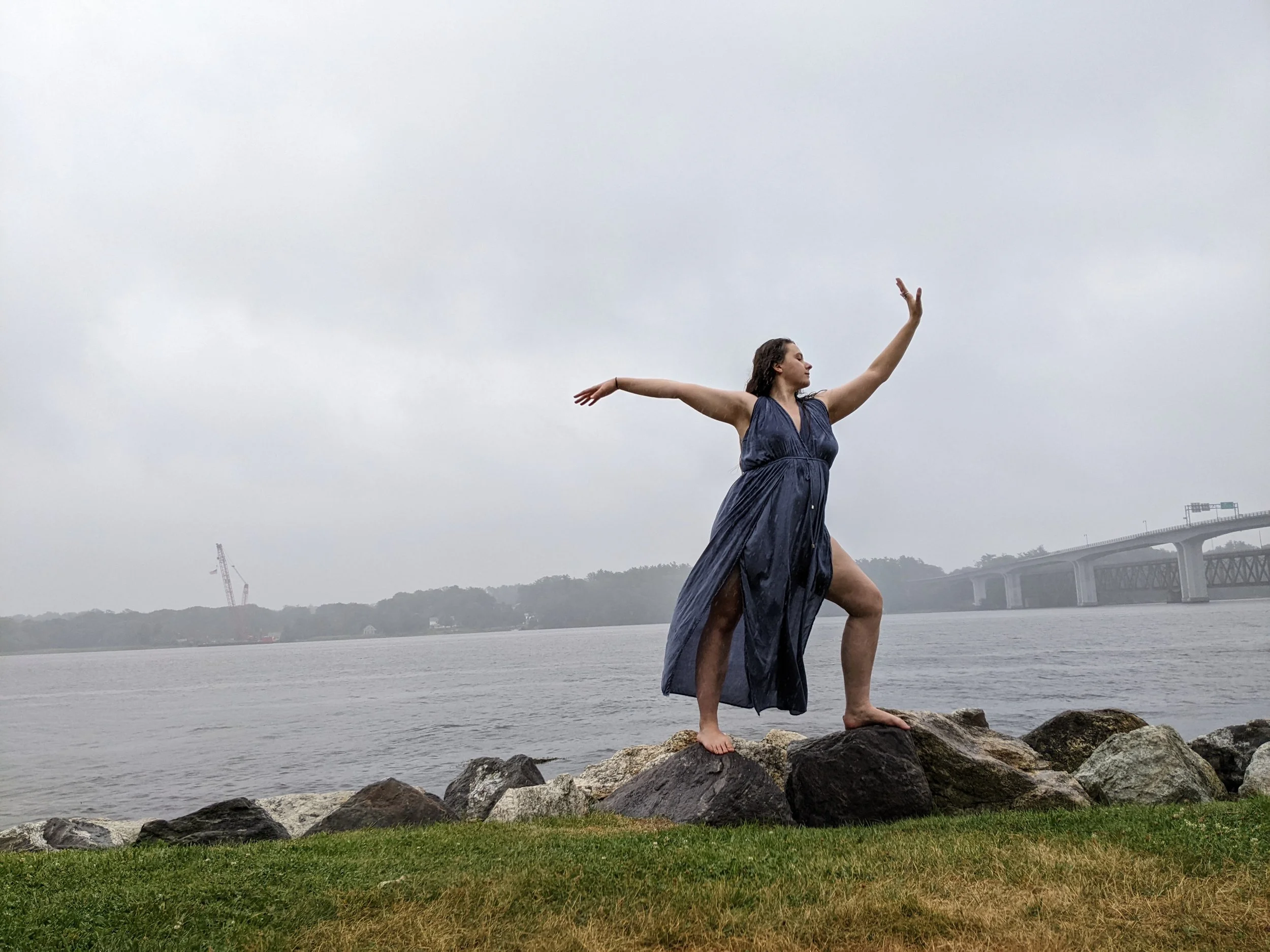 A woman in a flowy dress dances barefoot on rocks in the rain with a wide river behind her