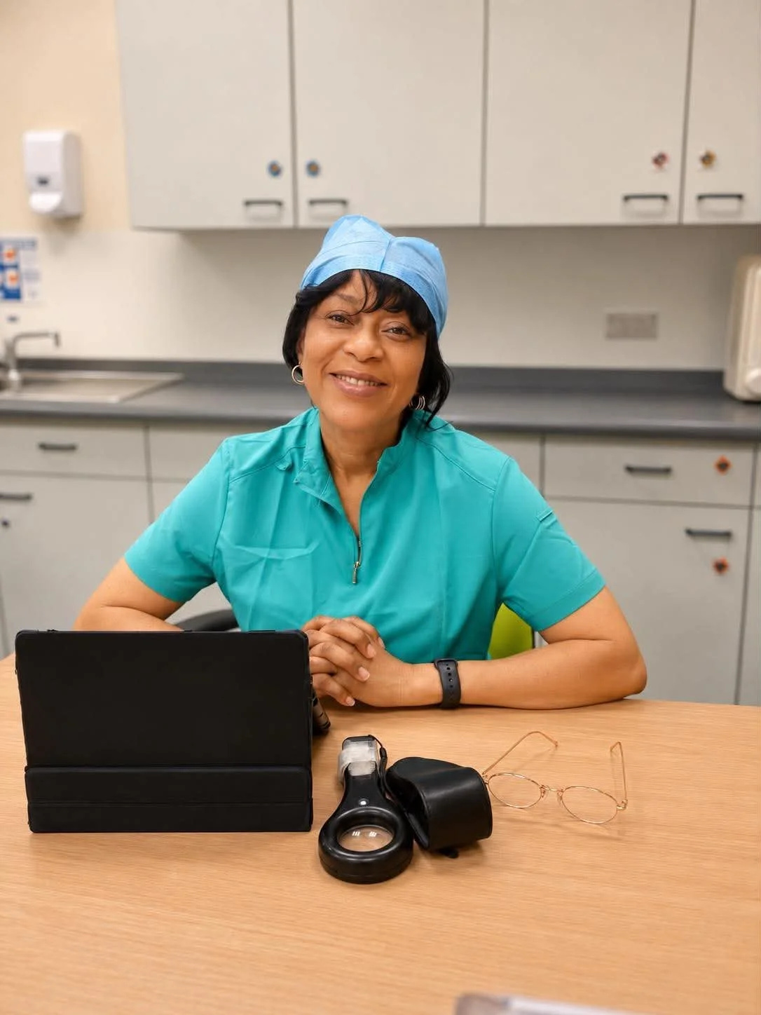 A female healthcare professional wearing a blue scrub cap and teal scrub top, sitting at a desk with a tablet, magnifying glass, and eyeglasses, in a medical office or hospital break room.